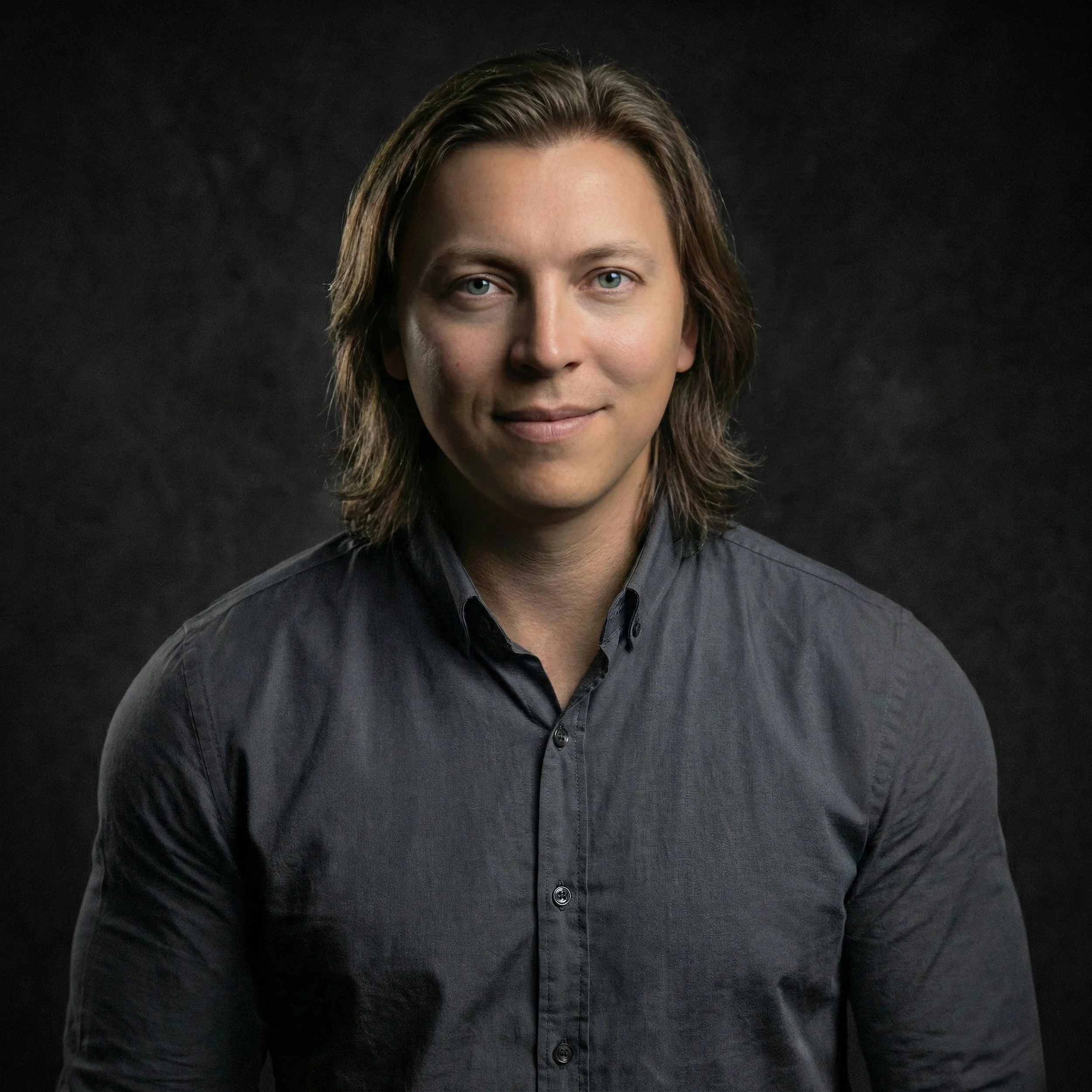 Close-up of a person's face with shoulder-length brown hair, blue eyes, light skin, wearing a dark button-up shirt, against a black background.