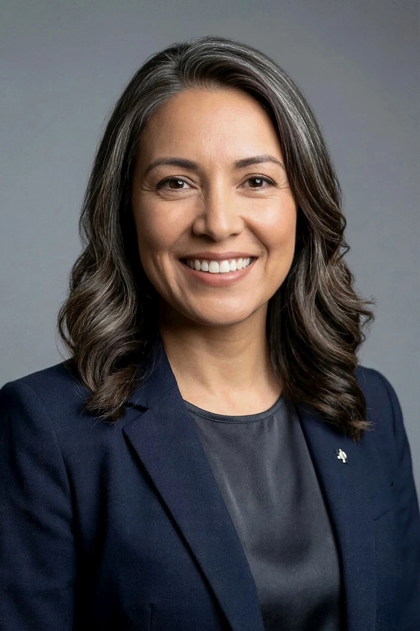 A woman with gray and brown wavy hair, wearing a dark blazer with a small pin, smiling against a plain background.