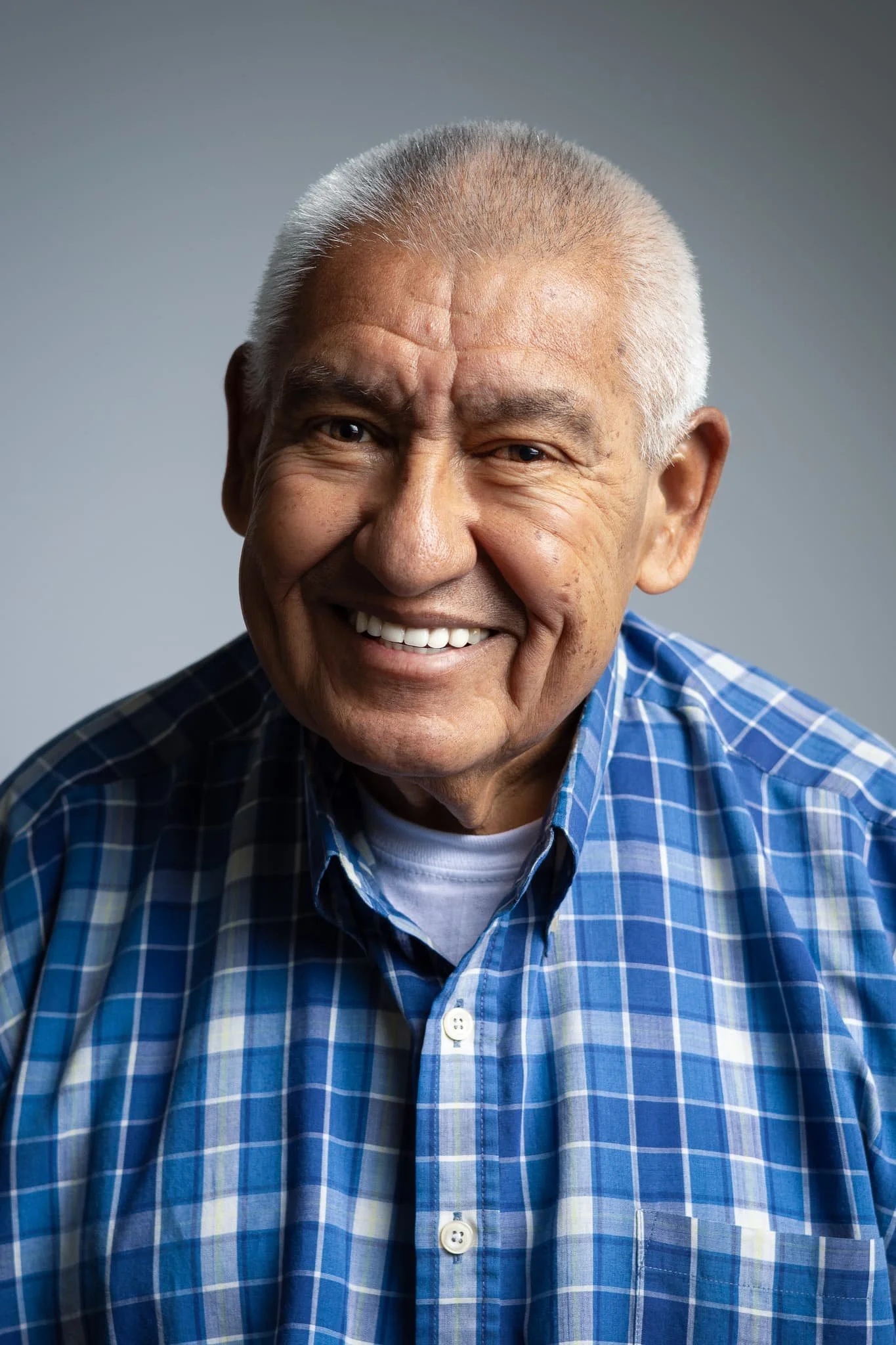 Close-up of smiling elderly man with short gray hair, wearing a blue plaid shirt.