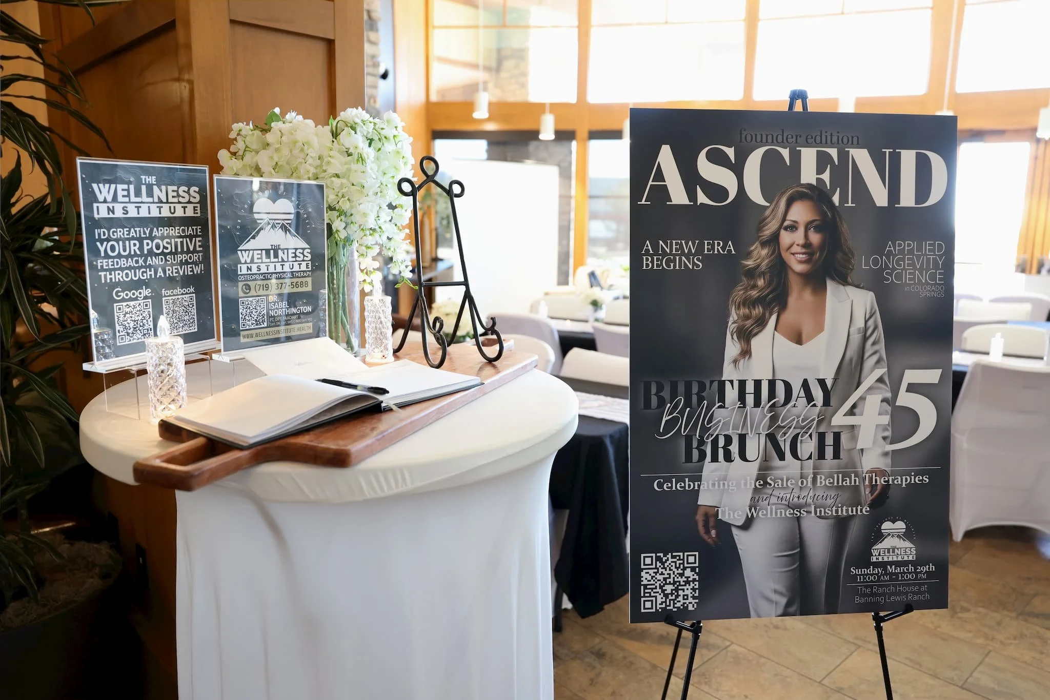 Event registration table featuring a floral arrangement, informational flyers about the Wellness Institute, and a promotional poster for a birthday brunch event with a smiling woman on it. The poster indicates the event is held at the Ranch House at 