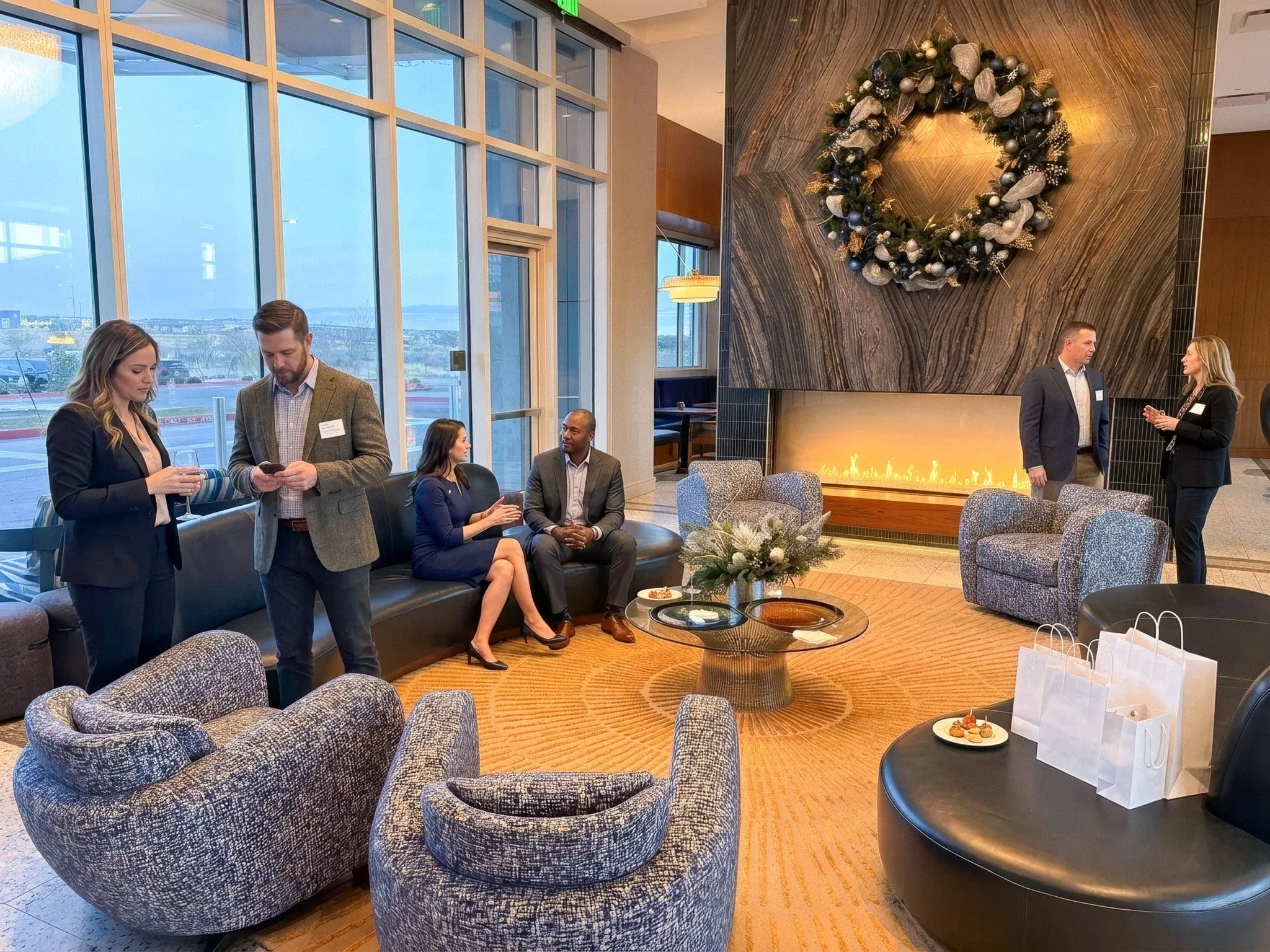 People gathered in a modern hotel lobby decorated for Christmas with a large wreath above a fireplace, and several groups of individuals engaged in conversation and using their phones, with cozy lounge chairs and a coffee table in the foreground.