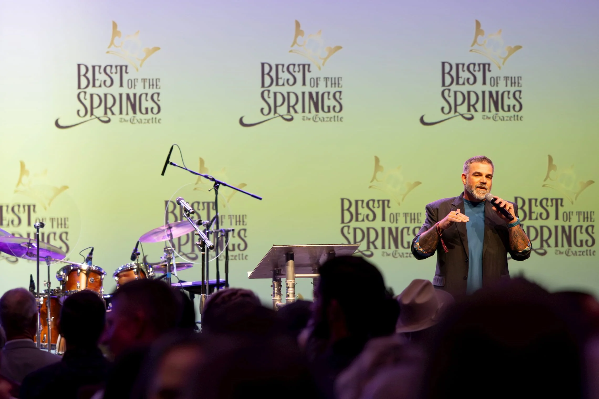 A man with gray hair and tattoos on his arms speaking into a microphone on stage at an event with a large backdrop that reads 'Best of the Springs The Gazette,' with musical instruments including drums set up nearby, and a partially visible audience 