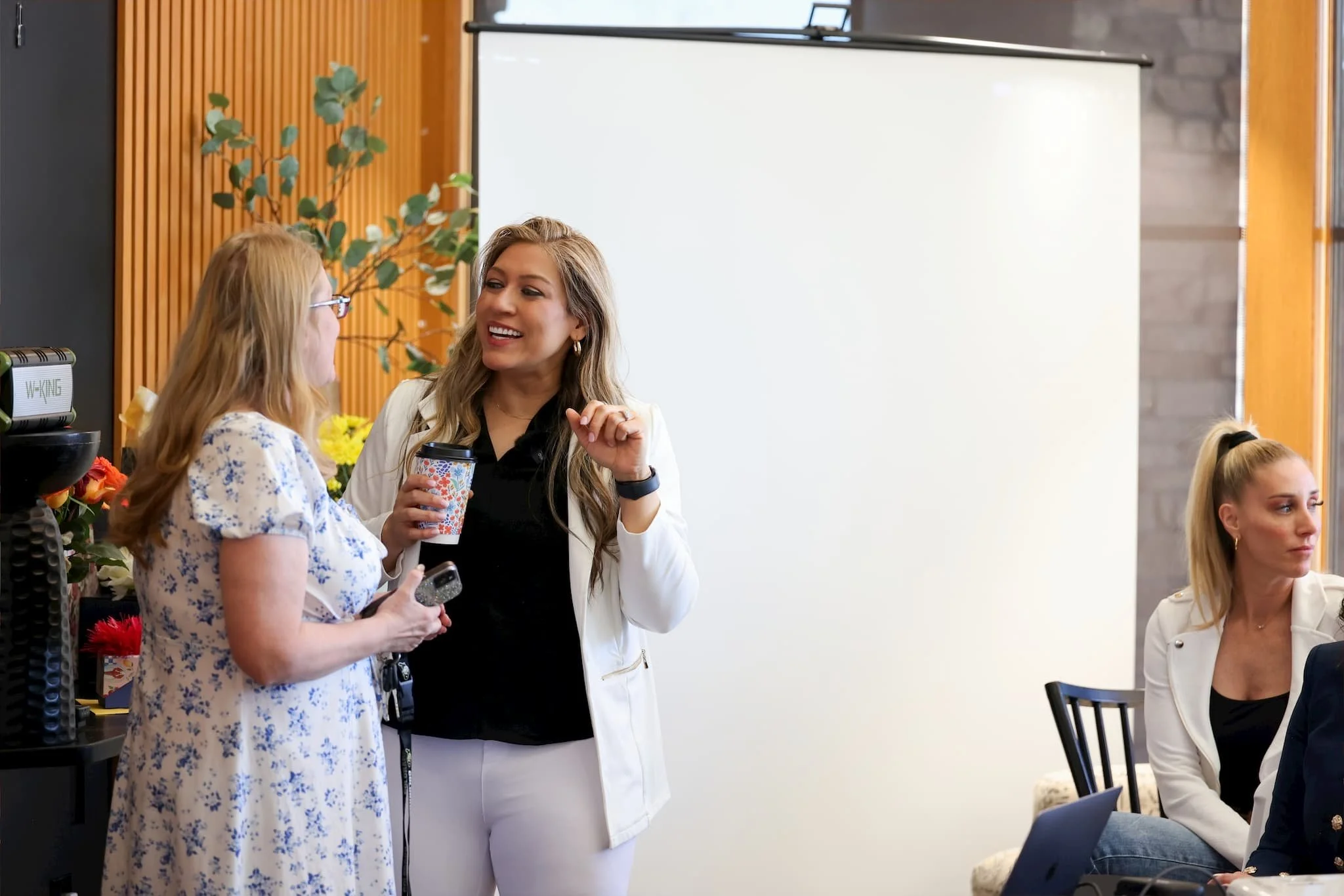 Two women are standing and engaging in a conversation, one is holding a coffee cup, and they are smiling. Other women are seated at a table in a room with wooden wall paneling and a whiteboard in the background.