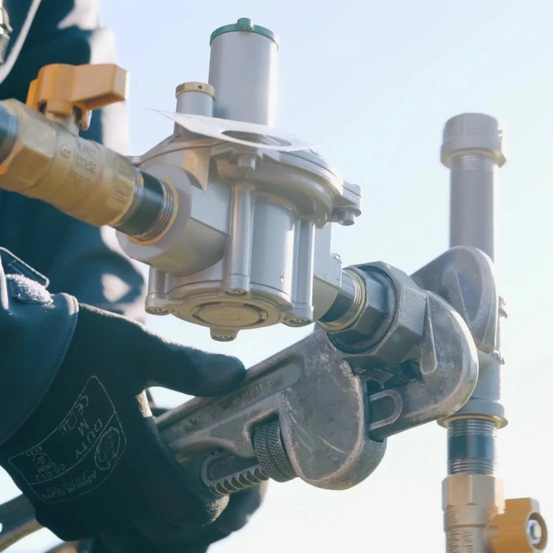 Close-up of a technician's hand in a black glove adjusting plumbing or gas equipment outdoors against a clear sky.