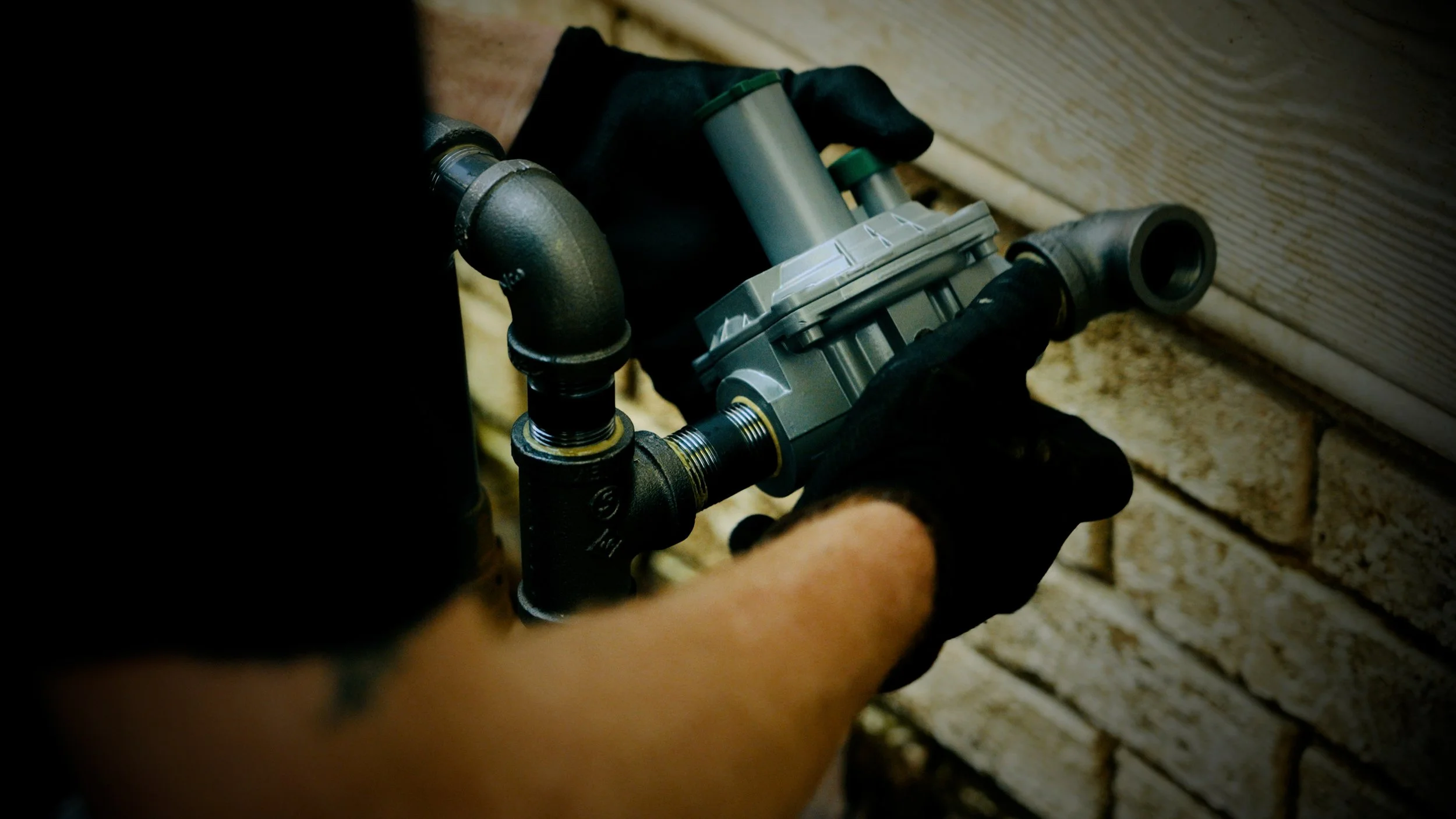 A person wearing black gloves is working with a pipe wrench on a pressure regulator or a similar component connected to pipes on a brick wall.