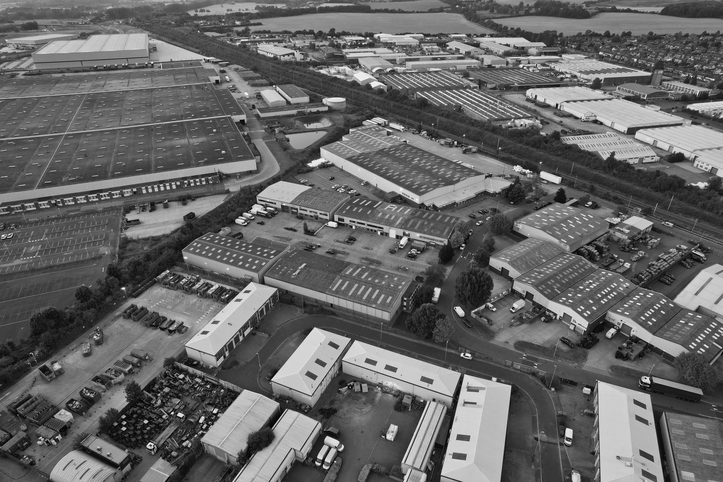 An aerial view of an industrial area with multiple warehouses, parking lots, and surrounding buildings. The landscape extends into fields and distant residential neighborhoods.