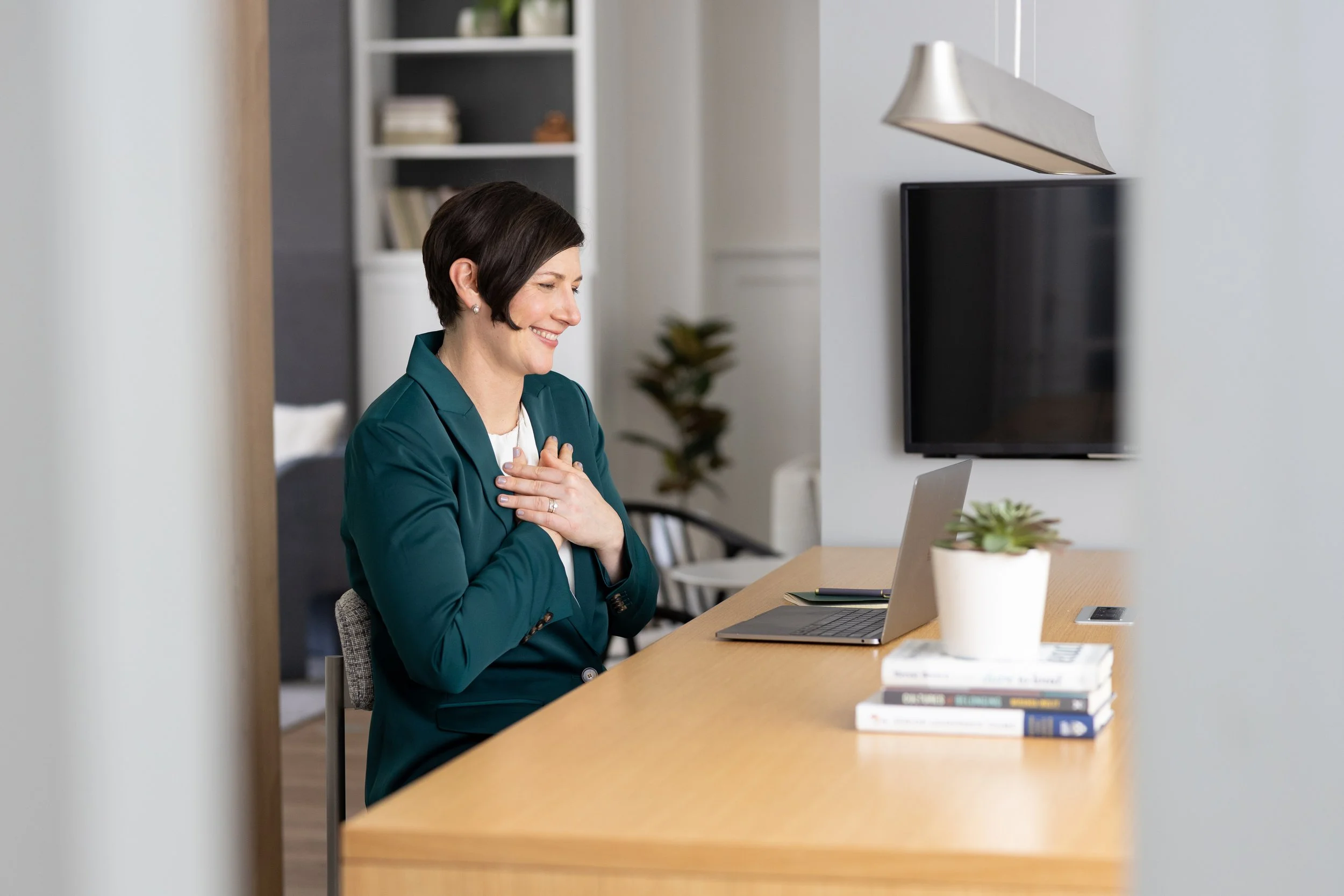 A woman with short dark hair smiling and placing her hand on her chest, sitting at a wooden desk with a laptop and a potted plant, in a modern office setting.