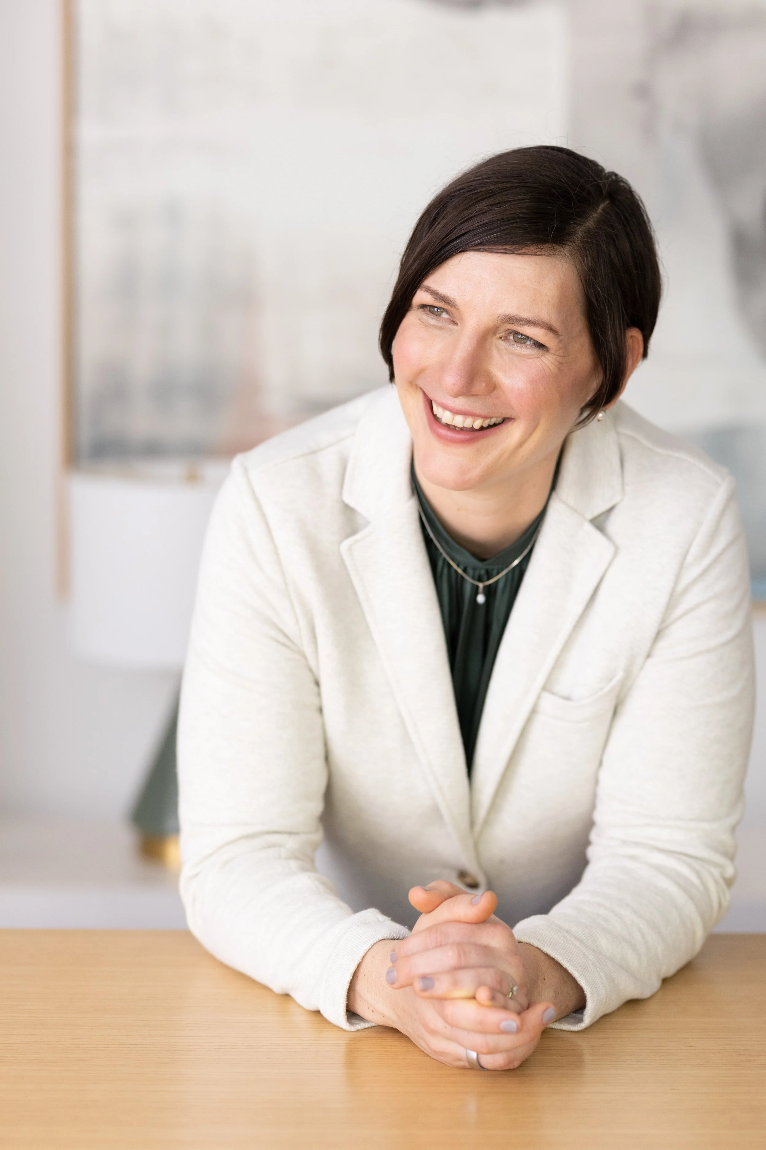 Woman with short dark hair smiling, sitting at a light wooden table, hands clasped, wearing a white blazer and black top.