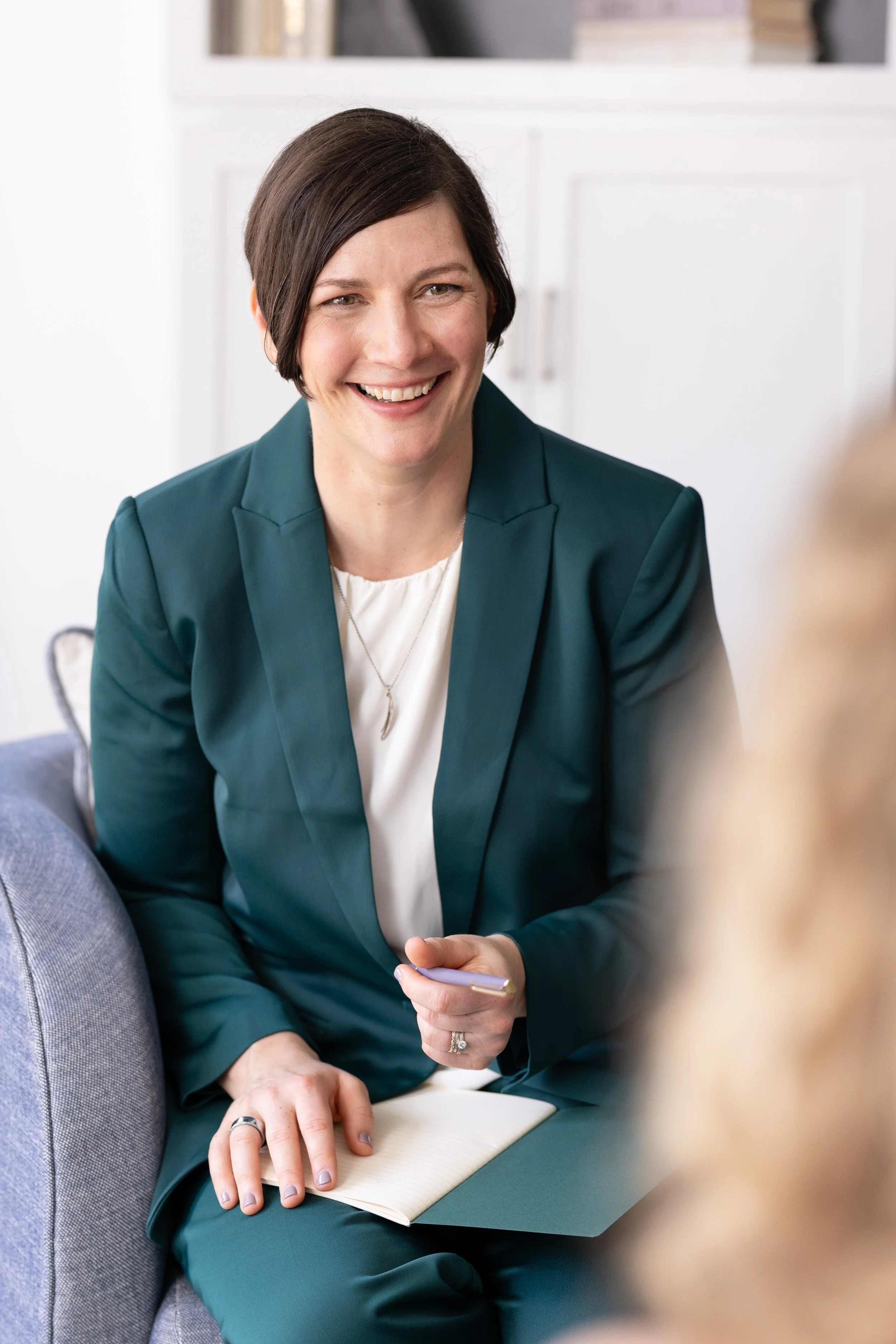 A woman with short dark hair smiling, wearing a teal blazer over a white top, sitting on a gray couch in a modern office or living room, holding a notebook and a pen.