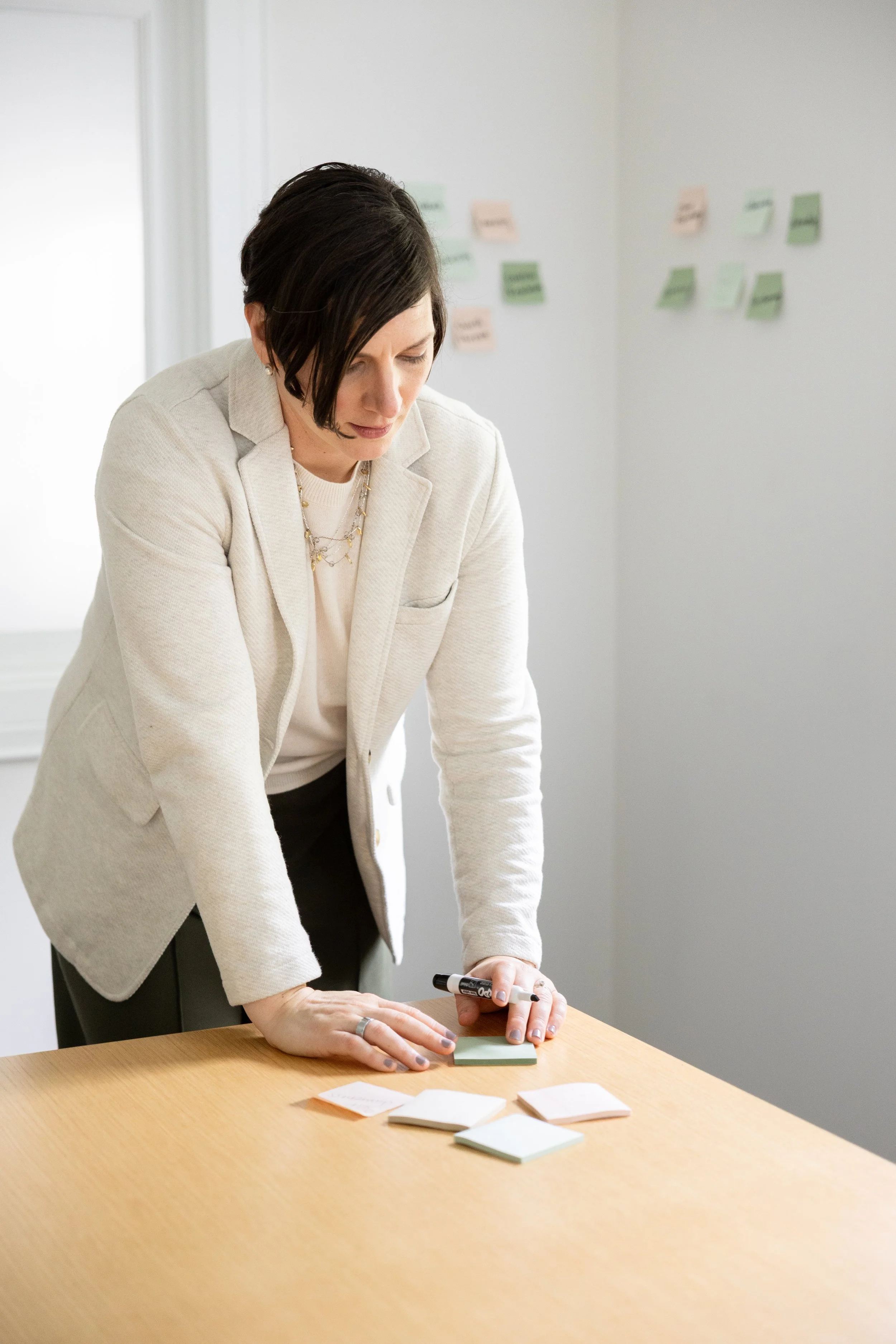 A woman in business attire is leaning over a table, writing on sticky notes with a marker. Several sticky notes are spread out on the table in front of her. The background shows a white wall with colorful sticky notes arranged in rows.