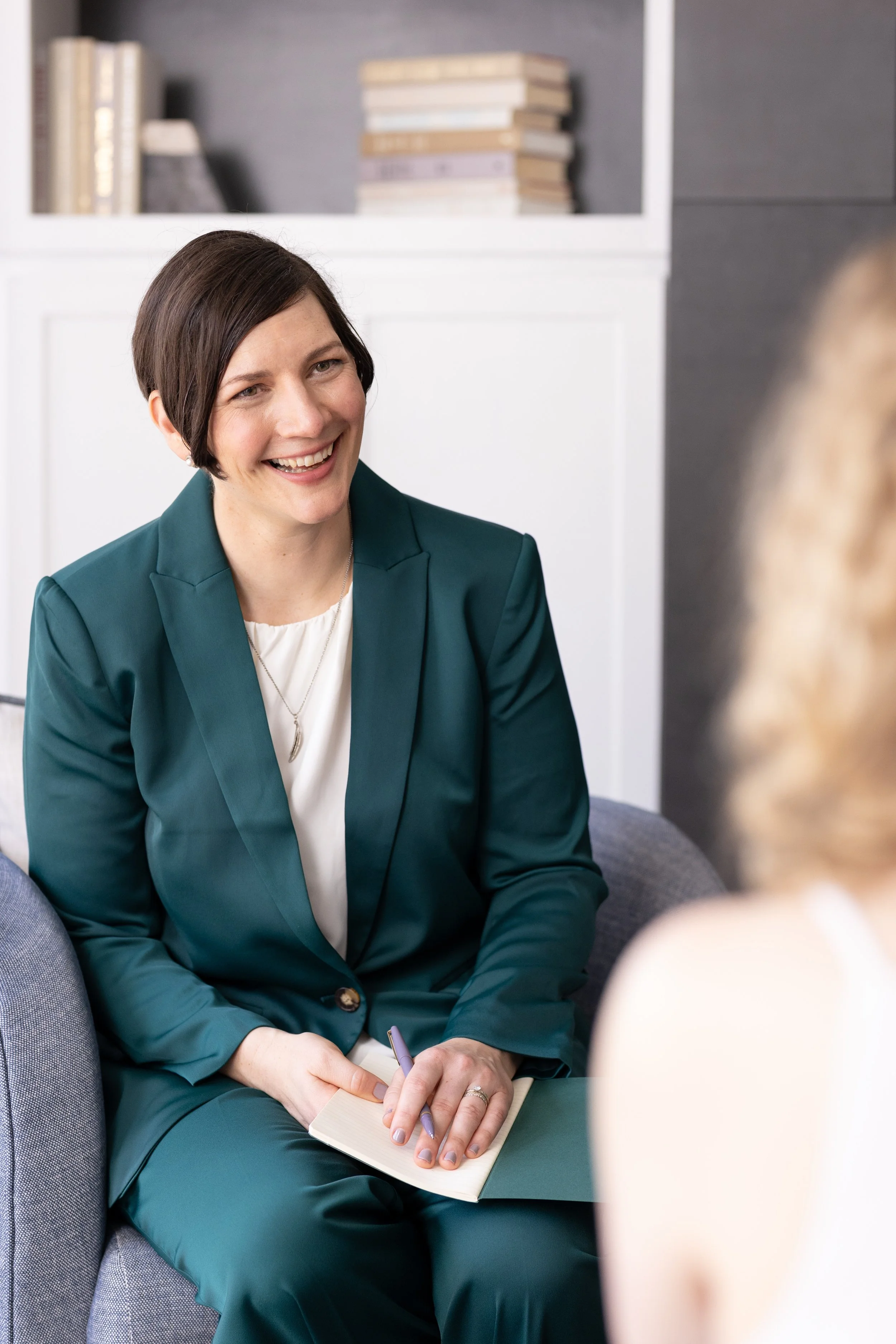 A woman with short dark hair, wearing a teal blazer and matching teal pants, smiling and talking with another person. She is sitting on a gray armchair, holding a notebook and pen. There are books on a shelf in the background.