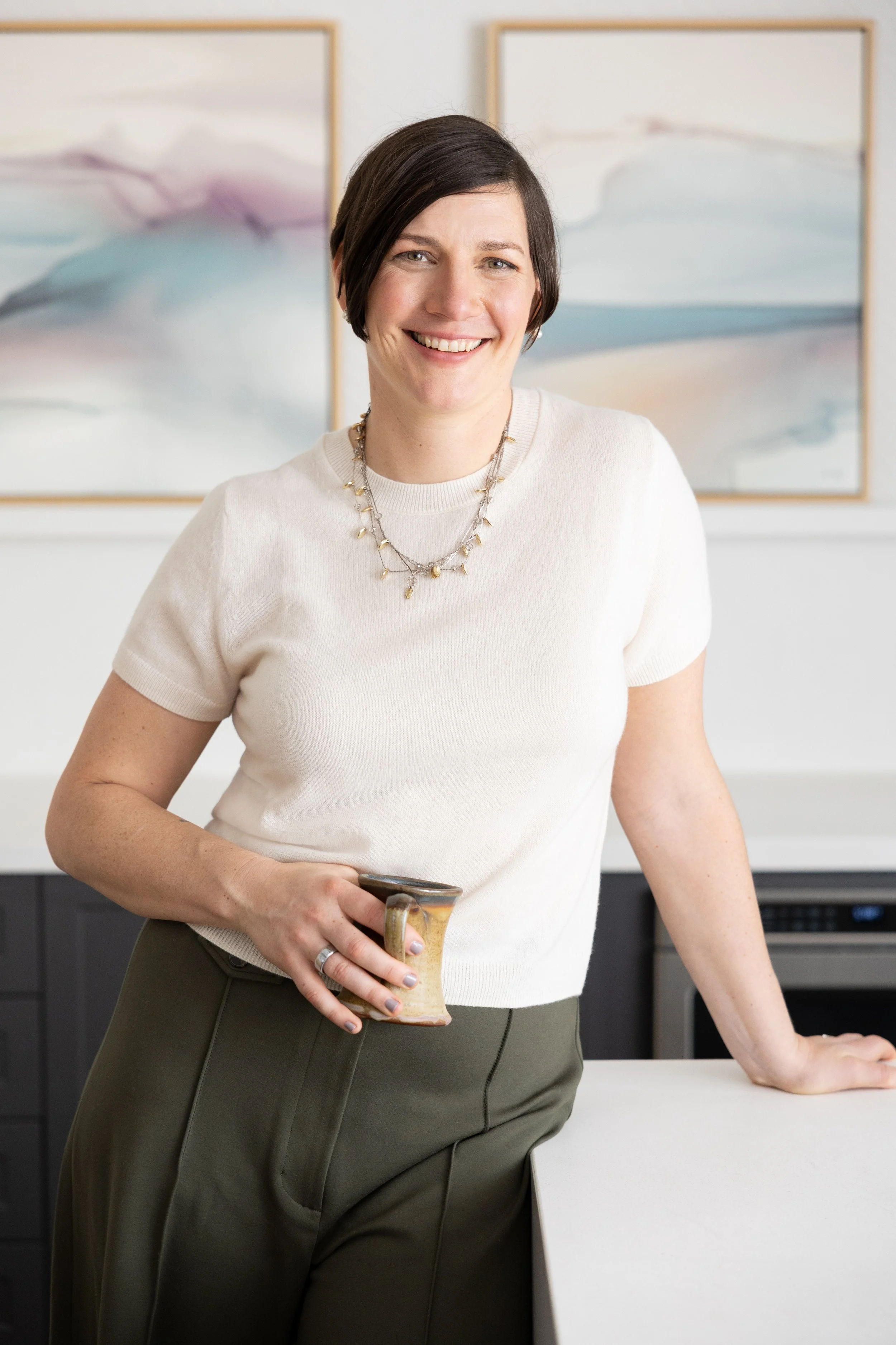 A woman with short dark hair smiling, wearing a white short-sleeved sweater and olive green pants, holding a ceramic coffee mug in her right hand, standing in a modern kitchen with abstract artwork on the wall behind her.