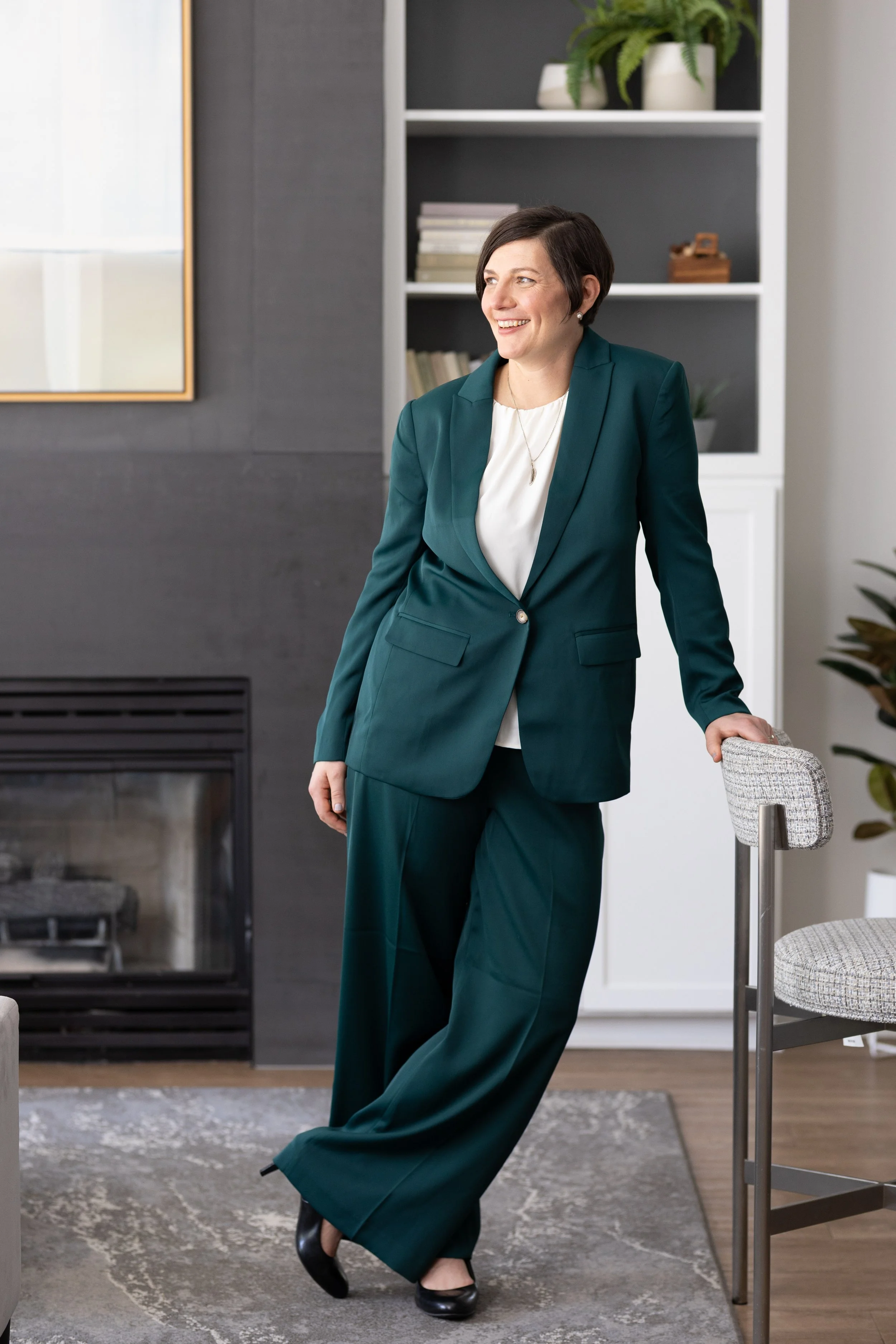 A woman with short dark hair, wearing a teal suit and black shoes, is leaning against a dining chair in a modern living room. There is a fireplace, a bookshelf with books, and potted plants in the background.