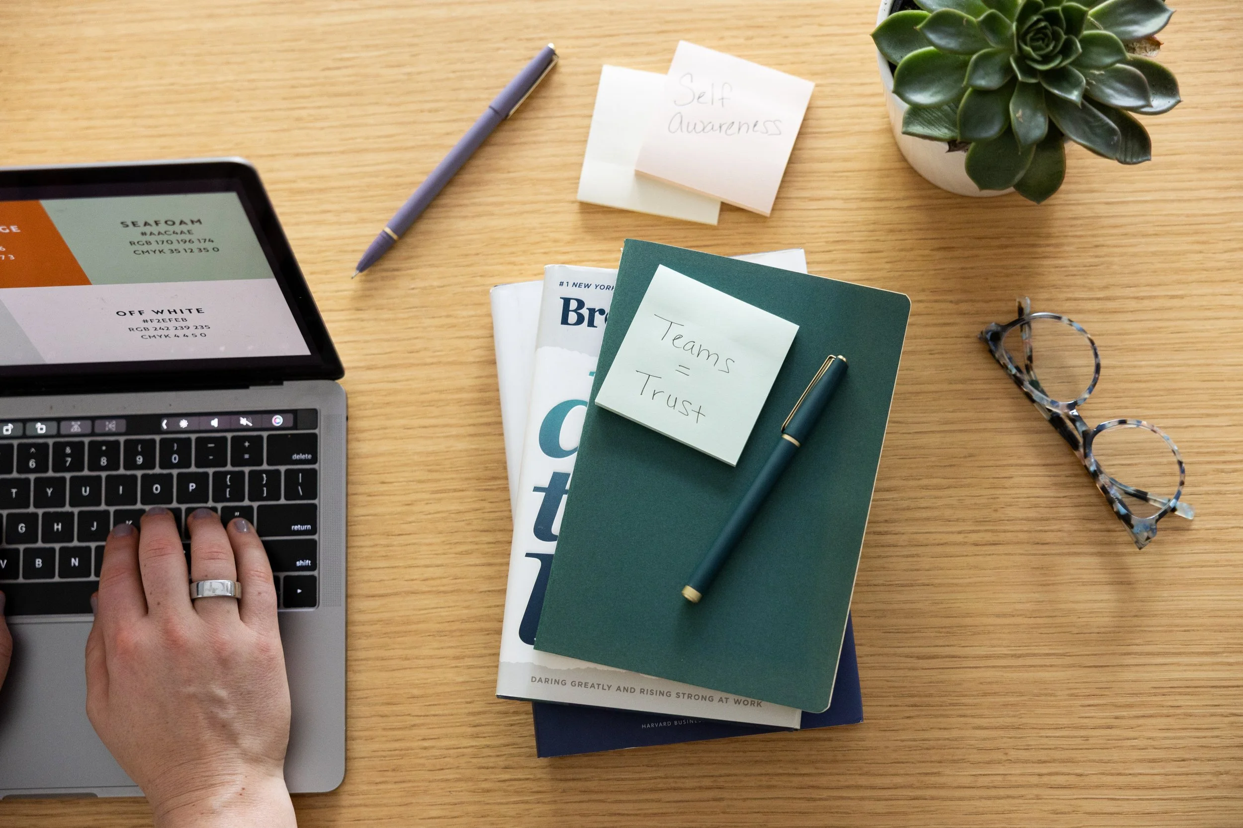 Wooden desk with a laptop, hand typing, notes with 'Self Awareness' and 'Teams = Trust', green succulent plant, eyeglasses, pen, and magazines.