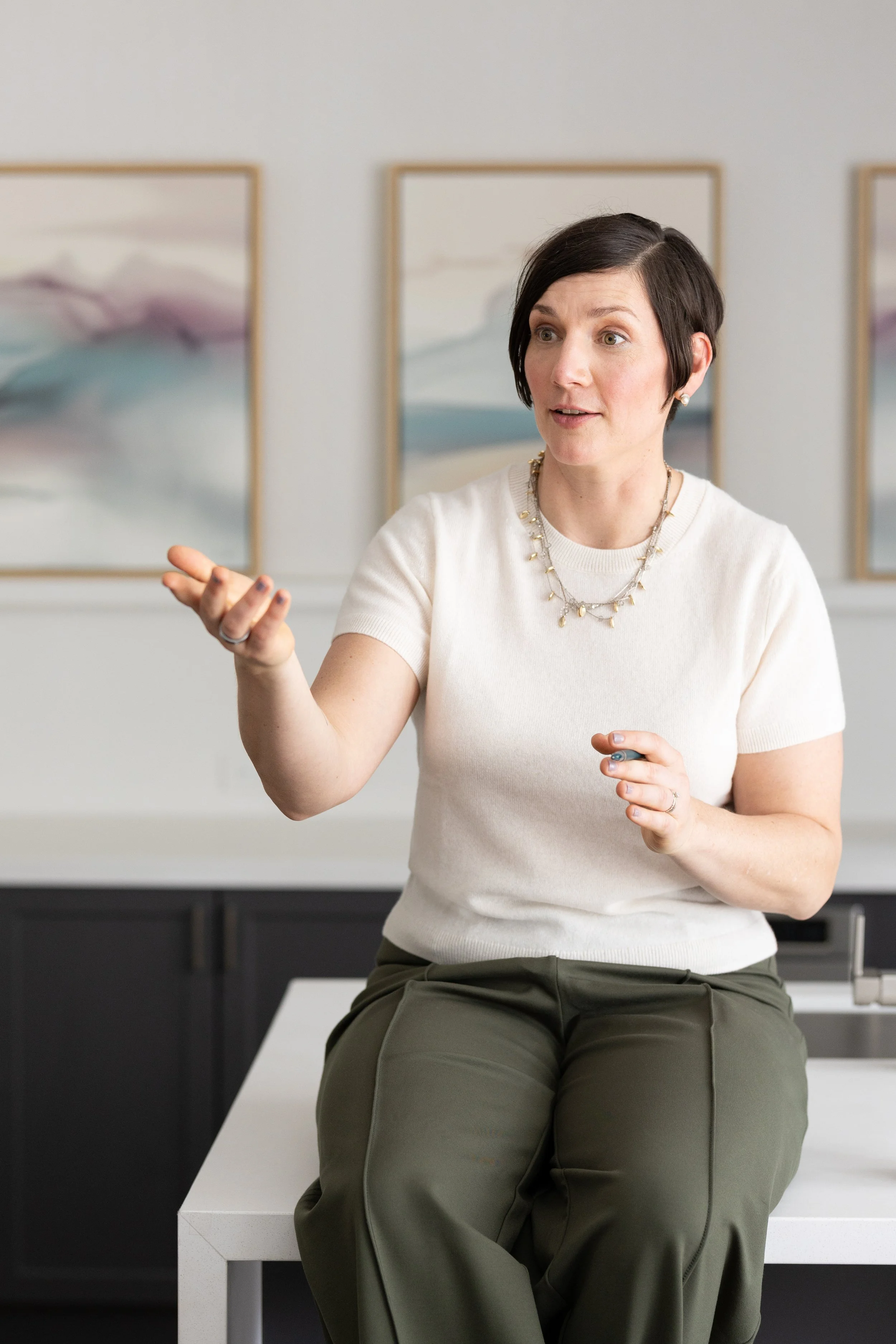 A woman with short dark hair, wearing a white shirt and olive green pants, sitting on a white countertop in a room with abstract artwork on the walls, gesturing with her hand while speaking.