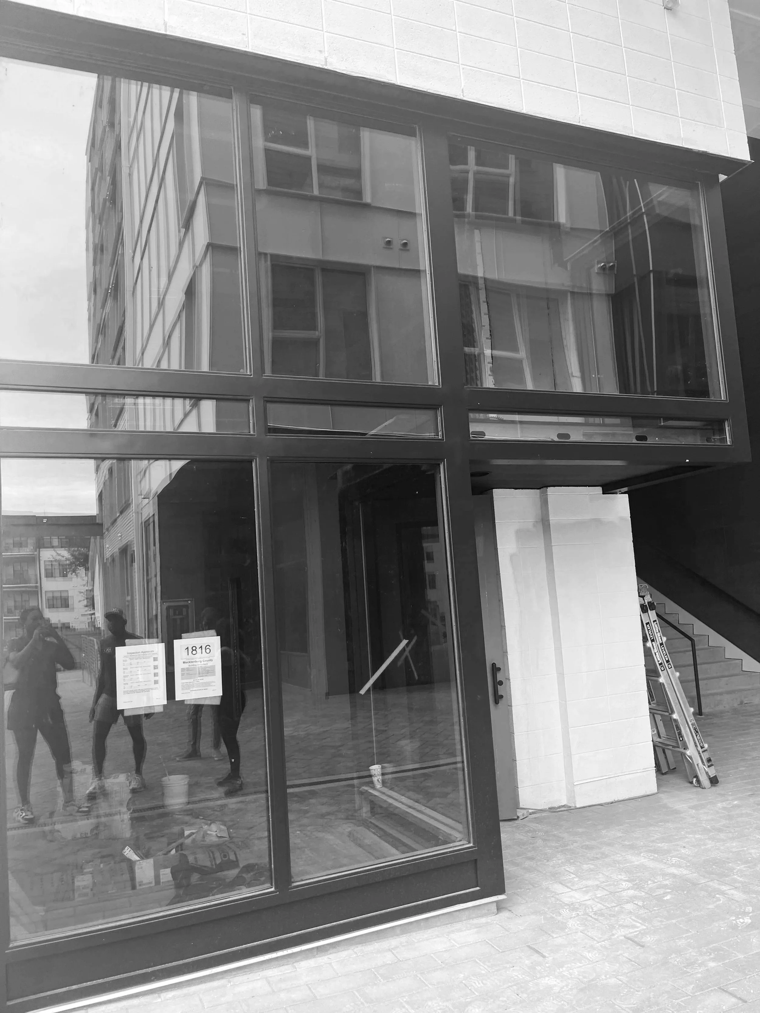 Construction site of a modern building with glass windows, reflection of people and surrounding buildings, opening in the front with signs and an aluminum ladder nearby.