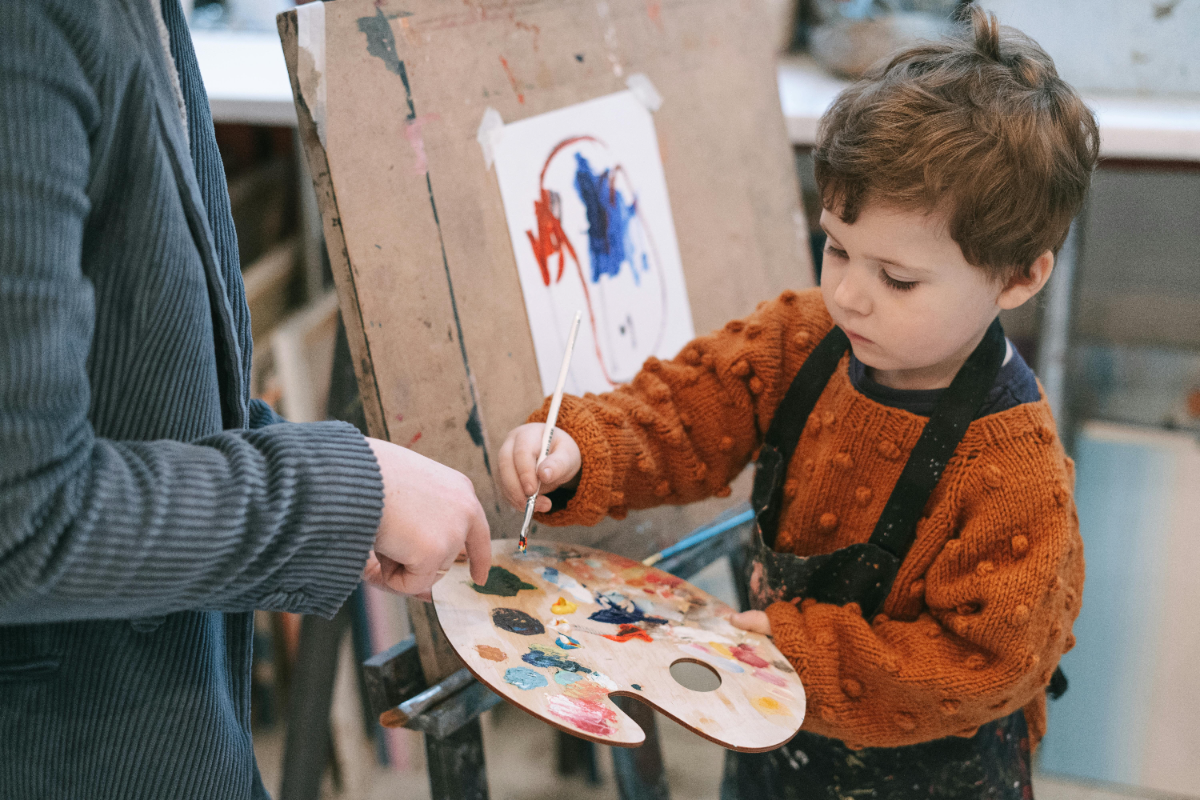 A young boy wearing an orange sweater and black apron is painting with guidance from an adult in an art studio. The boy holds a paintbrush and a color palette, focusing on his artwork on the easel.