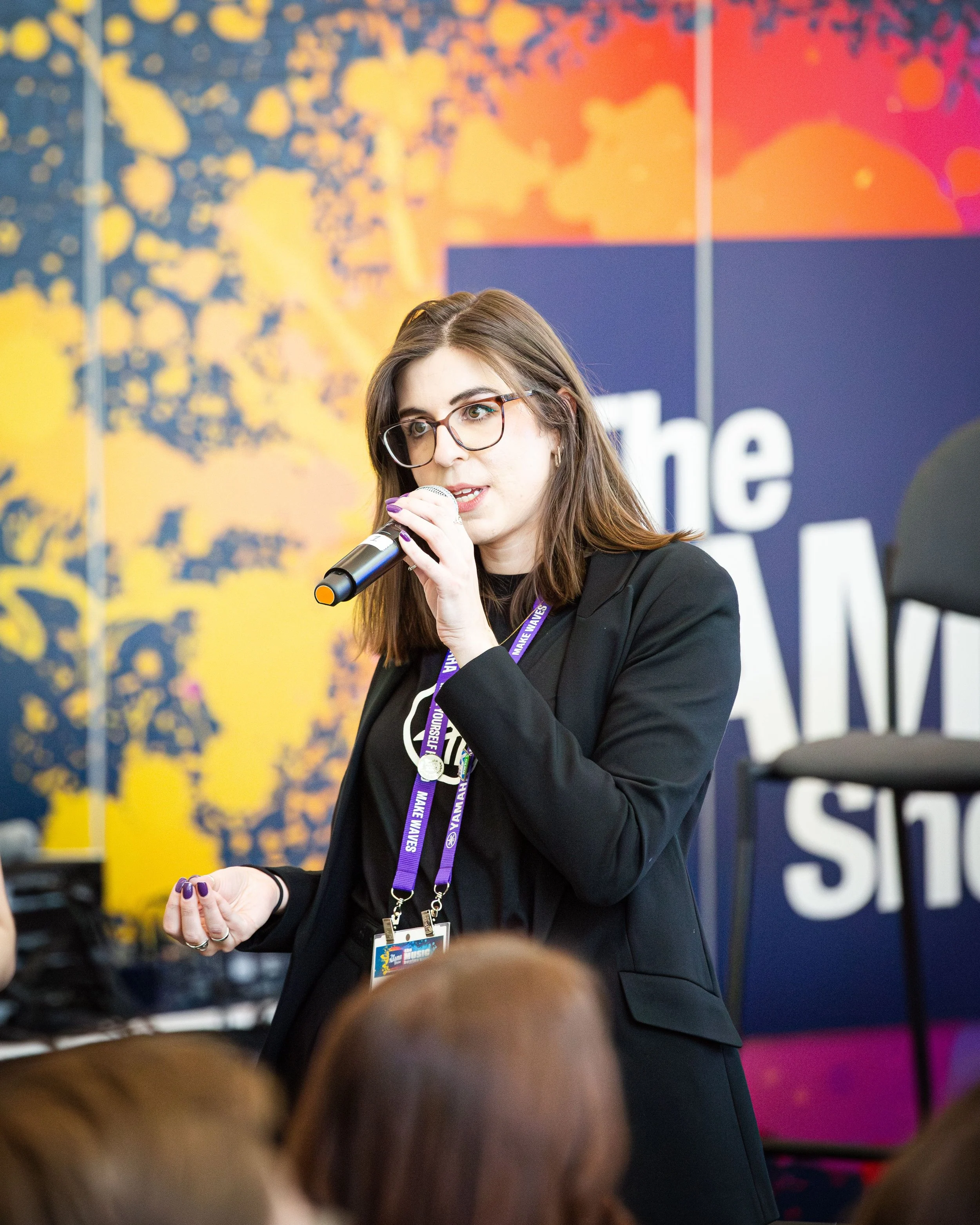 A young woman with glasses and brown hair speaking into a microphone at a conference, wearing a black blazer and lanyard.