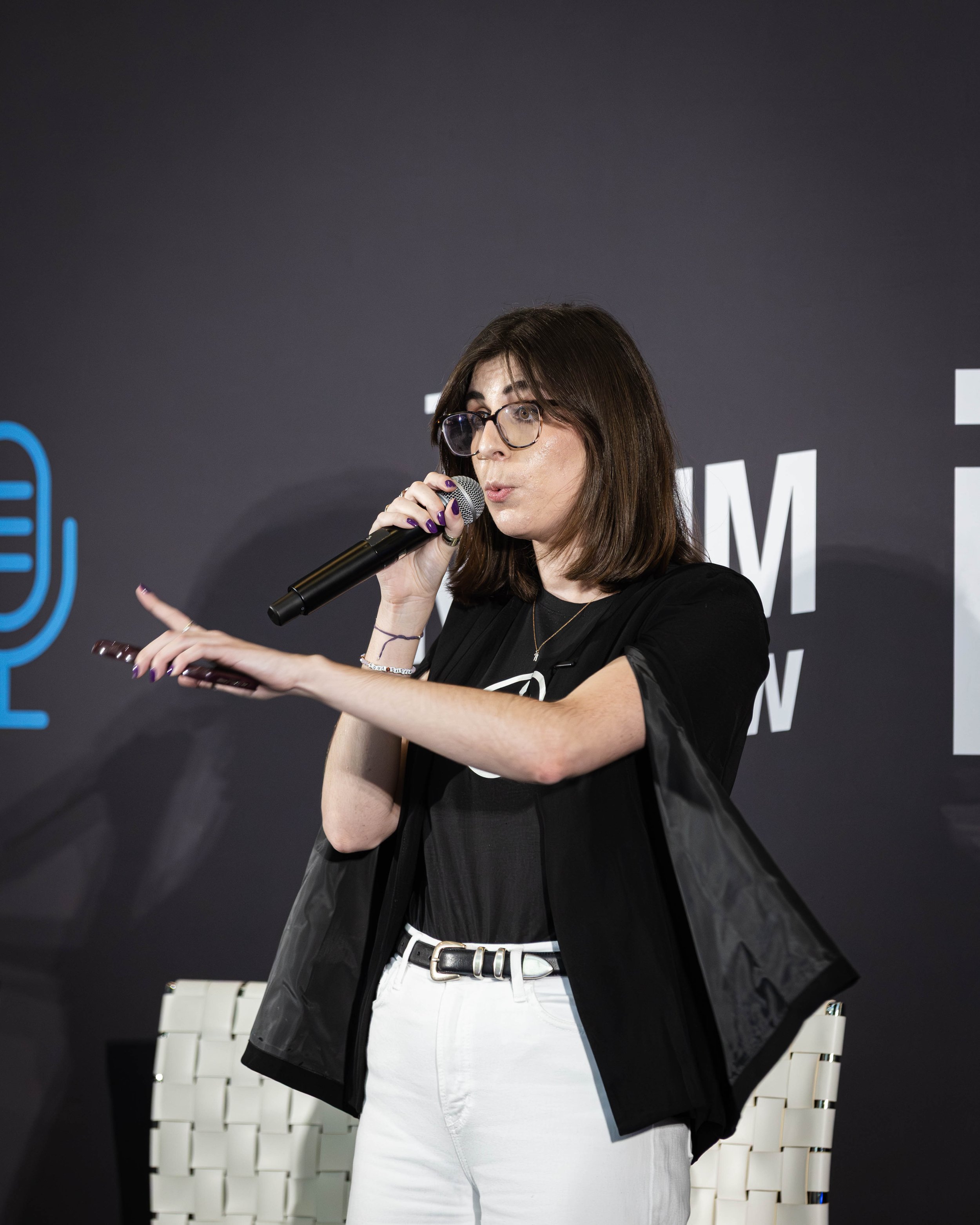 A woman with shoulder-length brown hair, glasses, wearing a black shirt and white pants, is holding a microphone and gesturing with her left hand while speaking on stage.