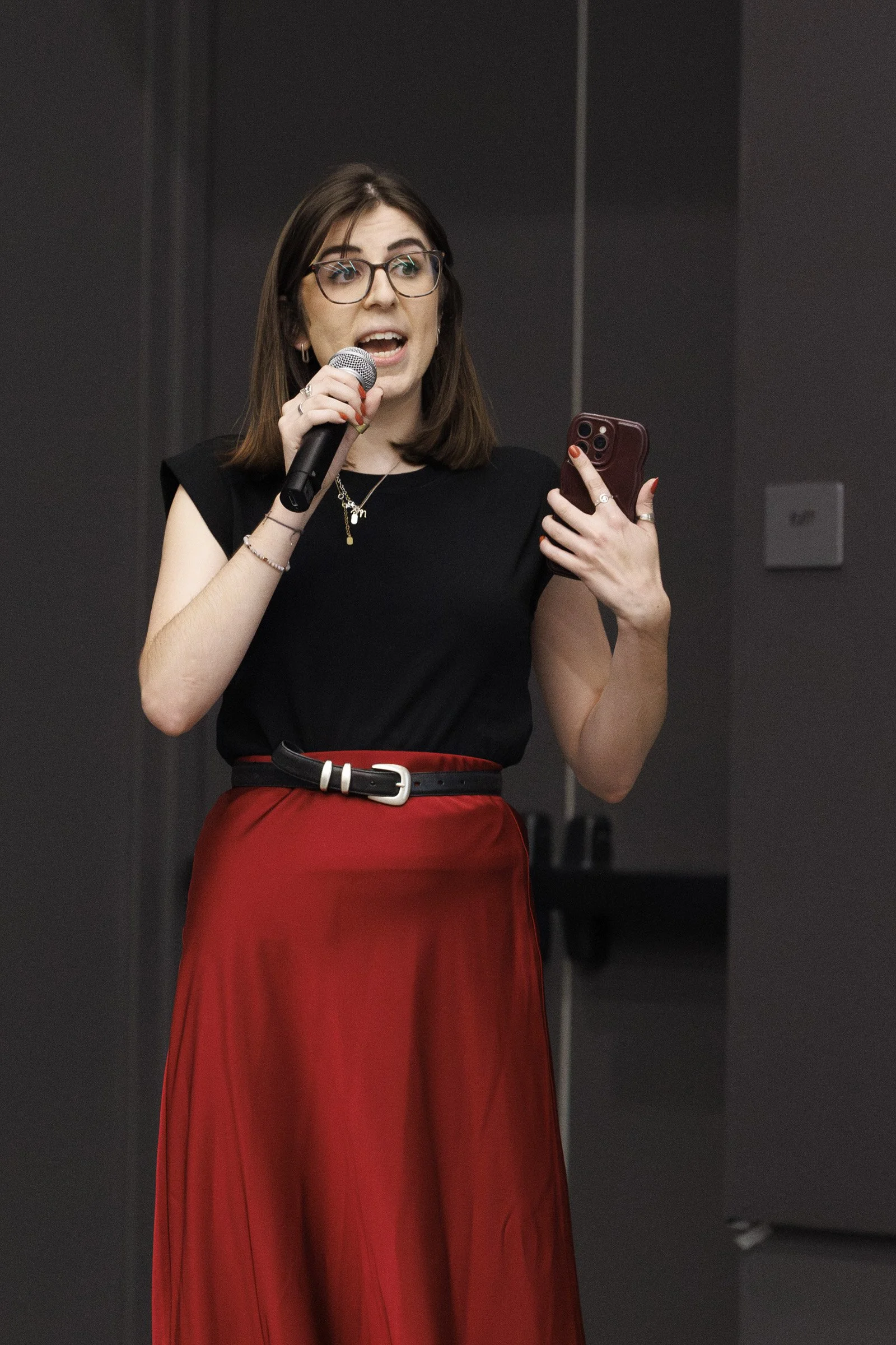 A young woman with glasses and brown hair, wearing a black top and a red skirt, holding a microphone in one hand and a phone in the other, speaking at an event.
