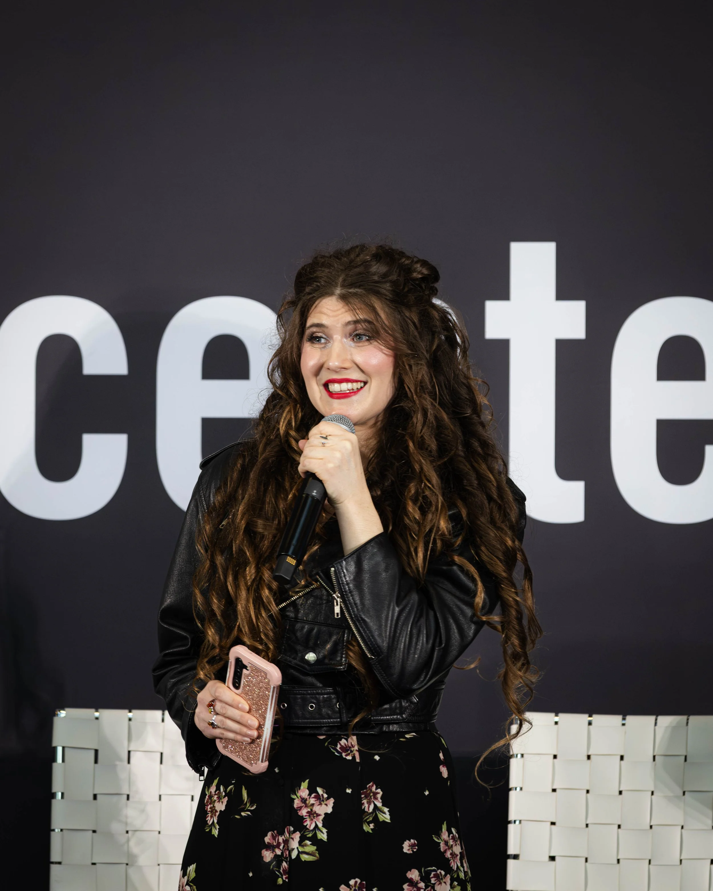 Woman with long curly hair speaking into a microphone, holding her phone, wearing a black leather jacket and floral skirt, standing in front of a black background with large white text.