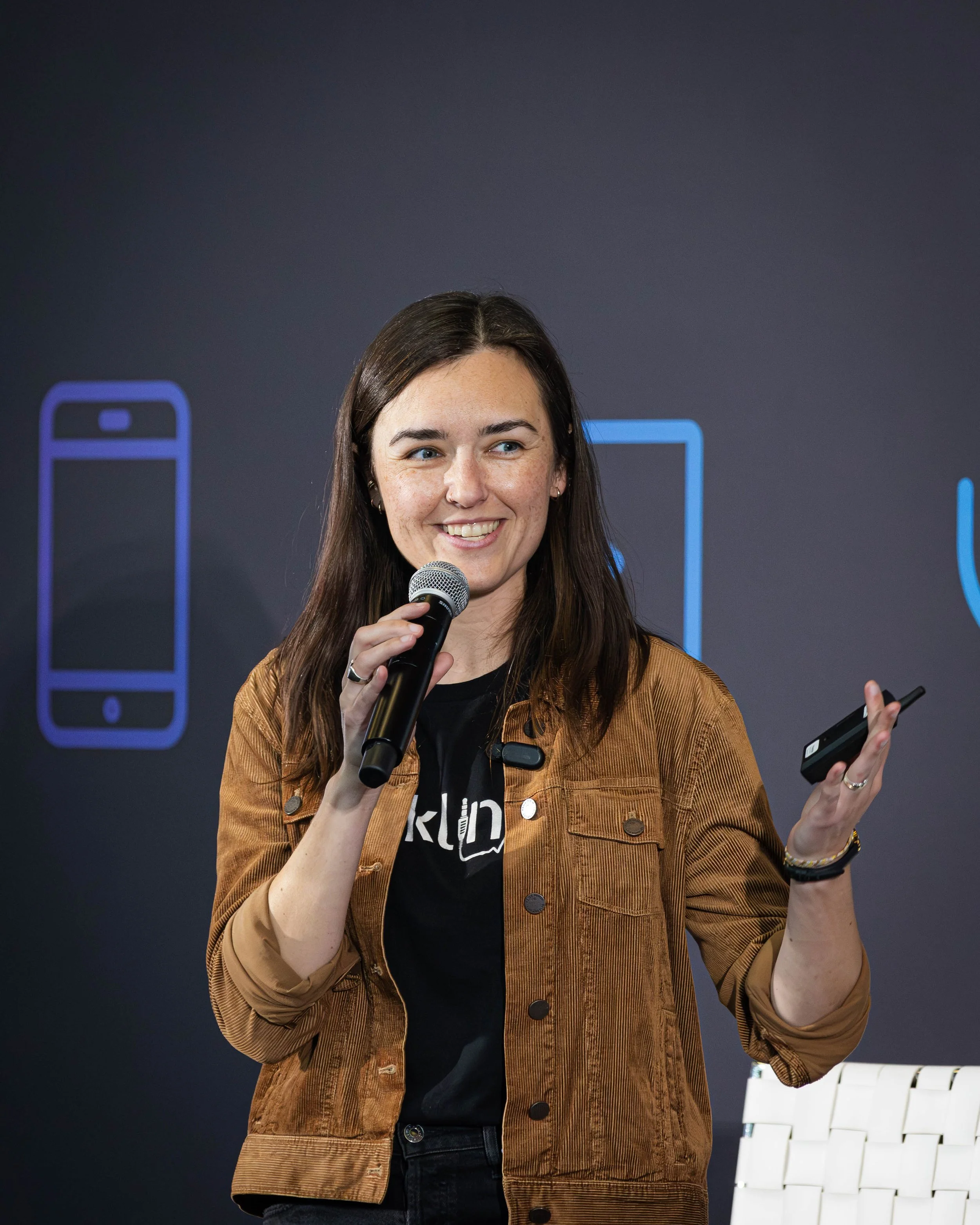 A woman with brown hair holding a microphone and a remote control, smiling and speaking at a presentation with illustrations of smartphones behind her.