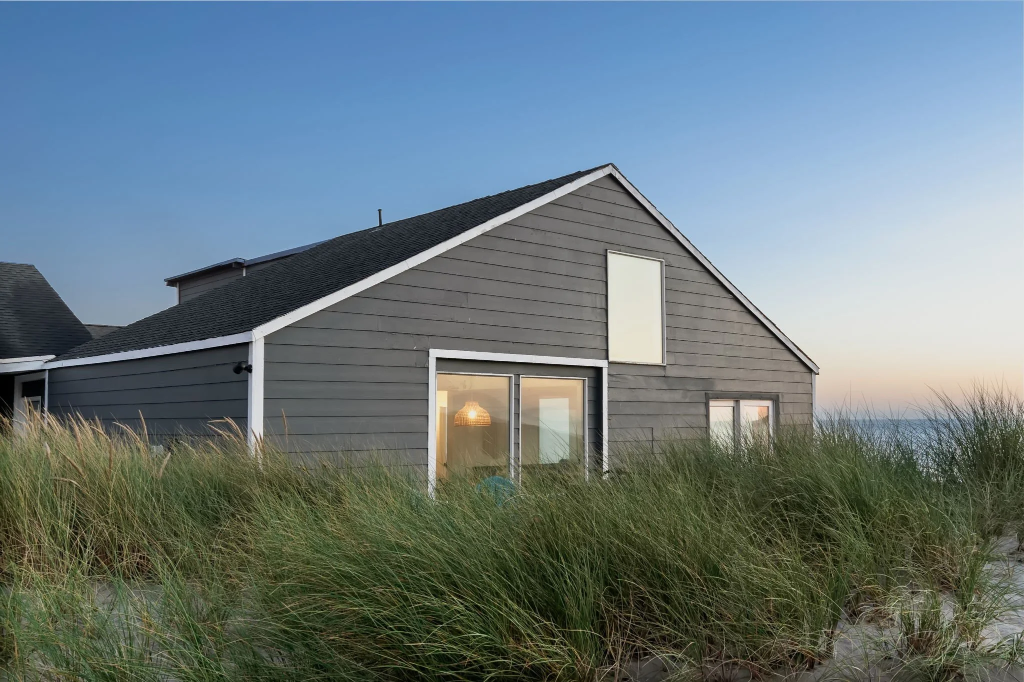 A gray house with a steep gabled roof, windows, and a door, surrounded by tall grass, under a clear blue sky during sunset.