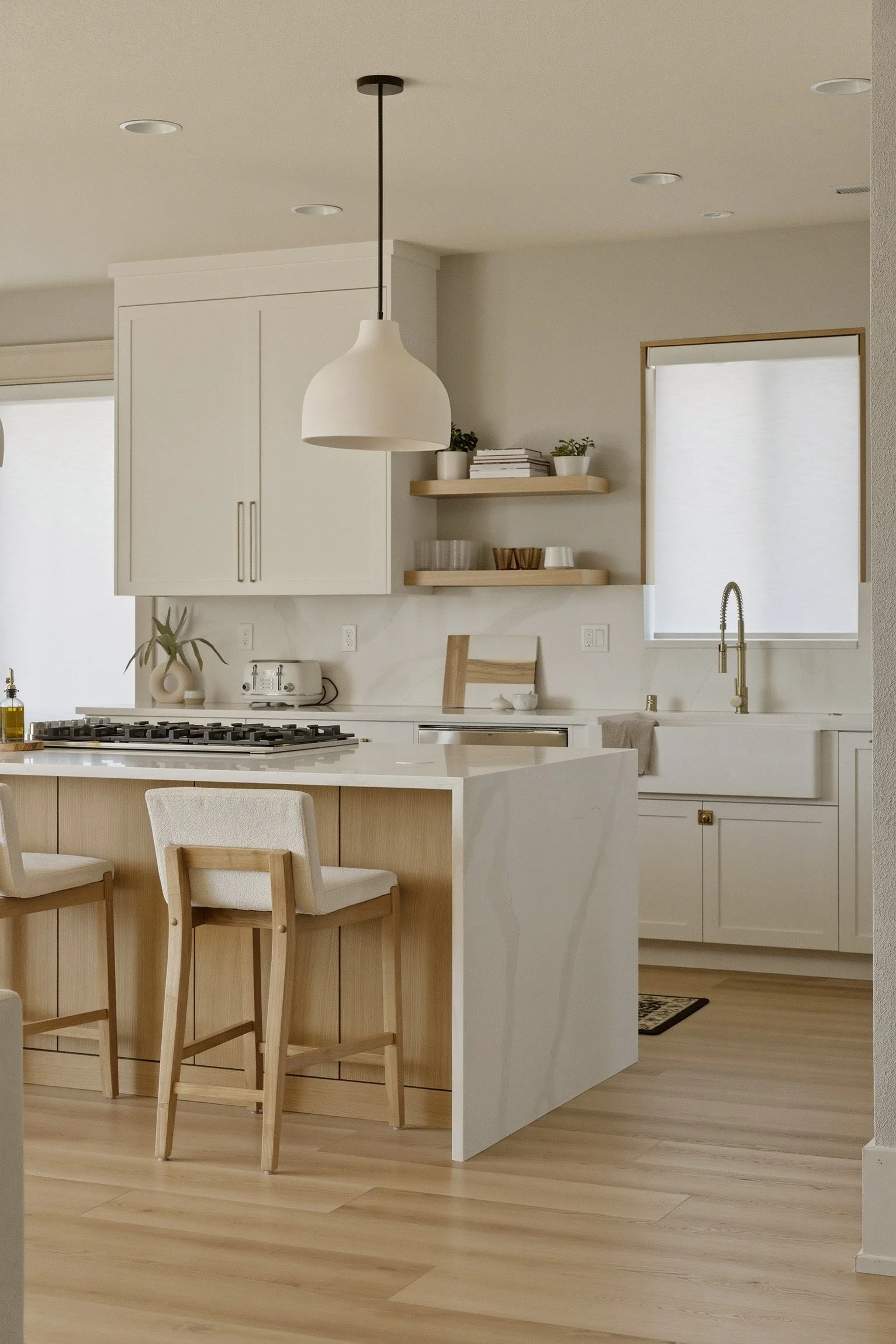 A modern kitchen with white cabinets, a marble countertop, a wooden island with two chairs, open shelving with potted plants and dishes, and a window with blinds. Toaster, plants, and a sink with a gold faucet are also visible.