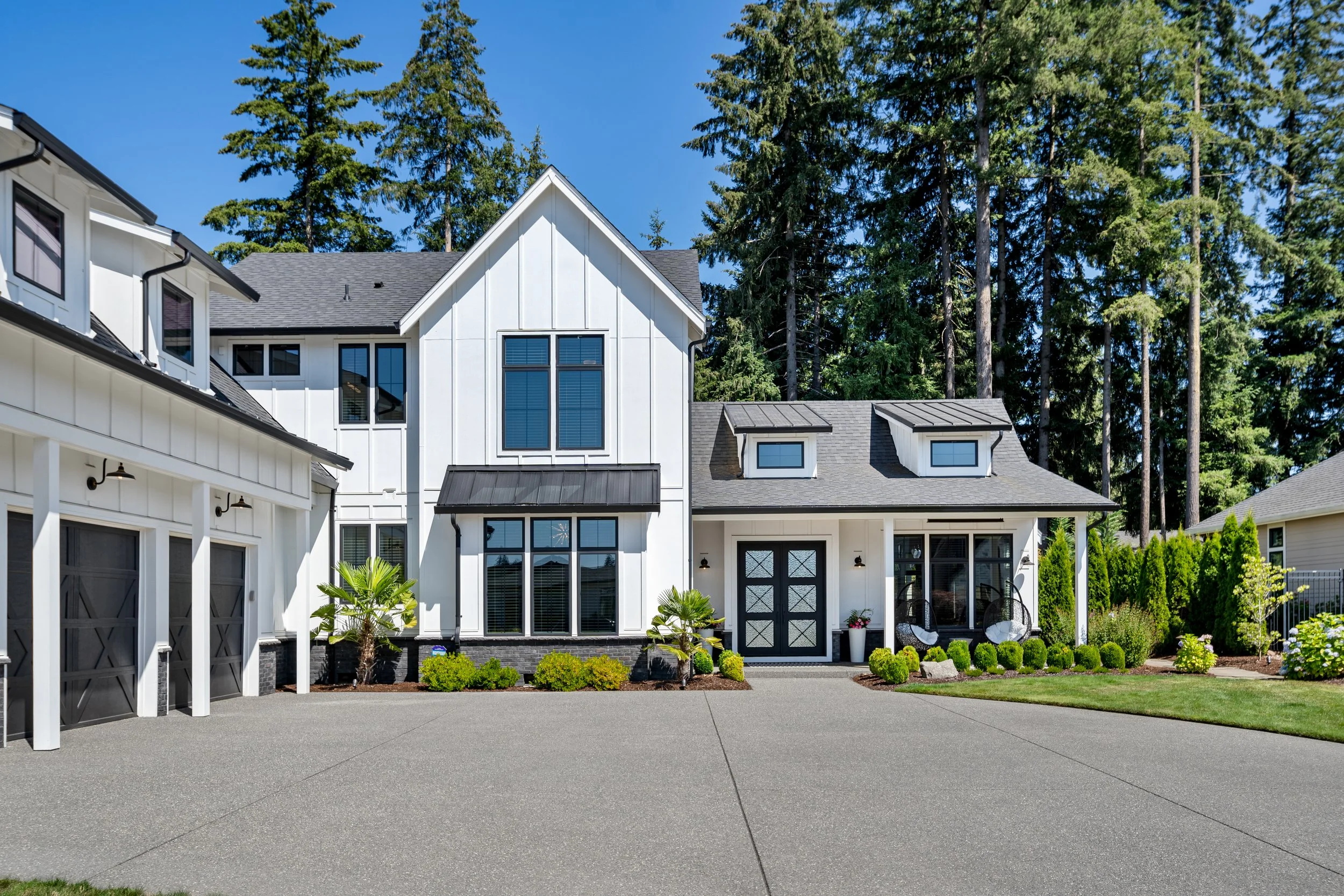 Modern white house with black accents, surrounded by greenery and tall trees, with a large driveway in the foreground.