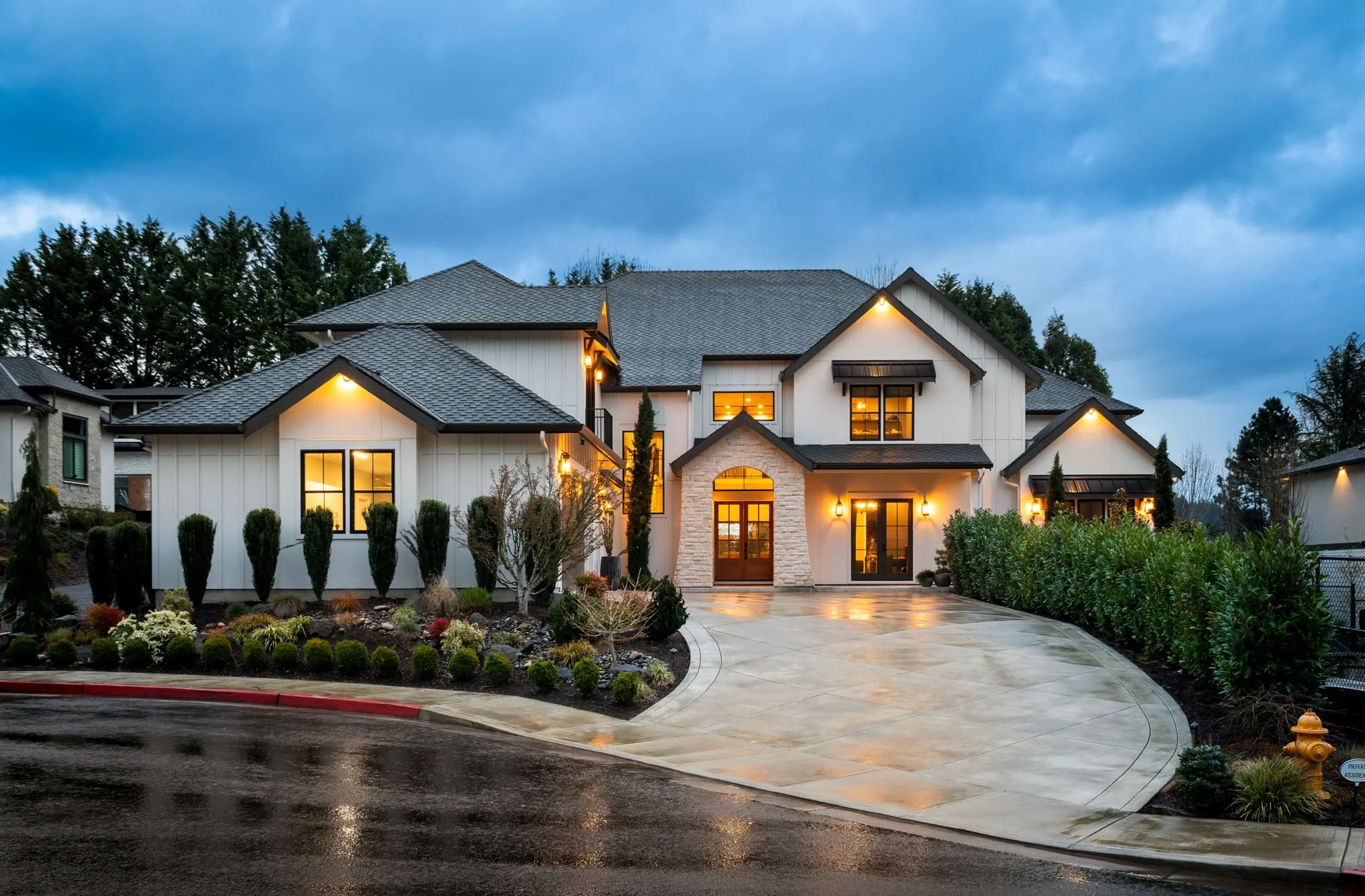 A modern two-story house with a stone and white exterior, multiple illuminated windows, and a landscaped front yard with bushes and small trees. The driveway is wet, and the sky is cloudy, suggesting evening or early night.