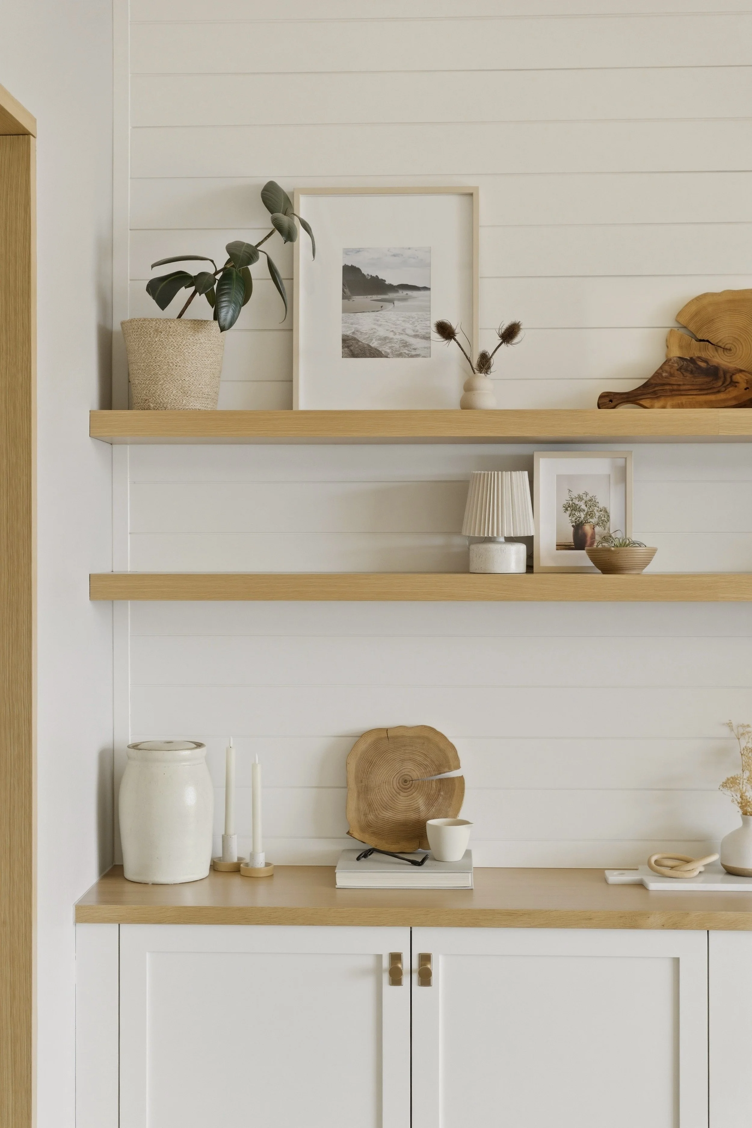 Minimalist interior with white shiplap wall and light wood shelves displaying decor such as framed beach photograph, potted plant, dried thistle flowers, pottery, books, and a decorative wood slice.