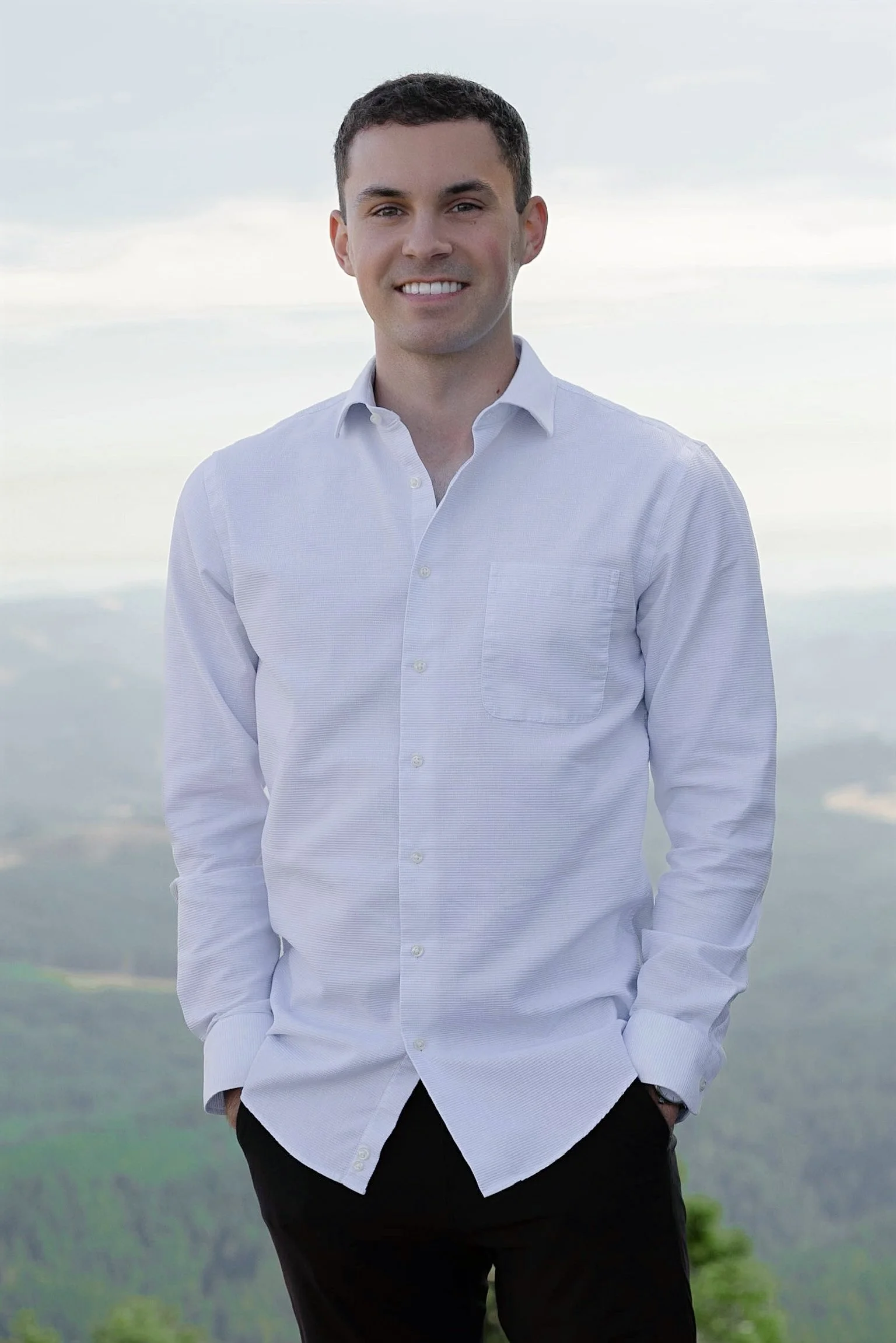 A young man with short dark hair, light skin, and a bright smile is standing outdoors against a backdrop of cloudy sky and mountains. He is wearing a white button-up shirt and dark pants, with his hand in his pocket.