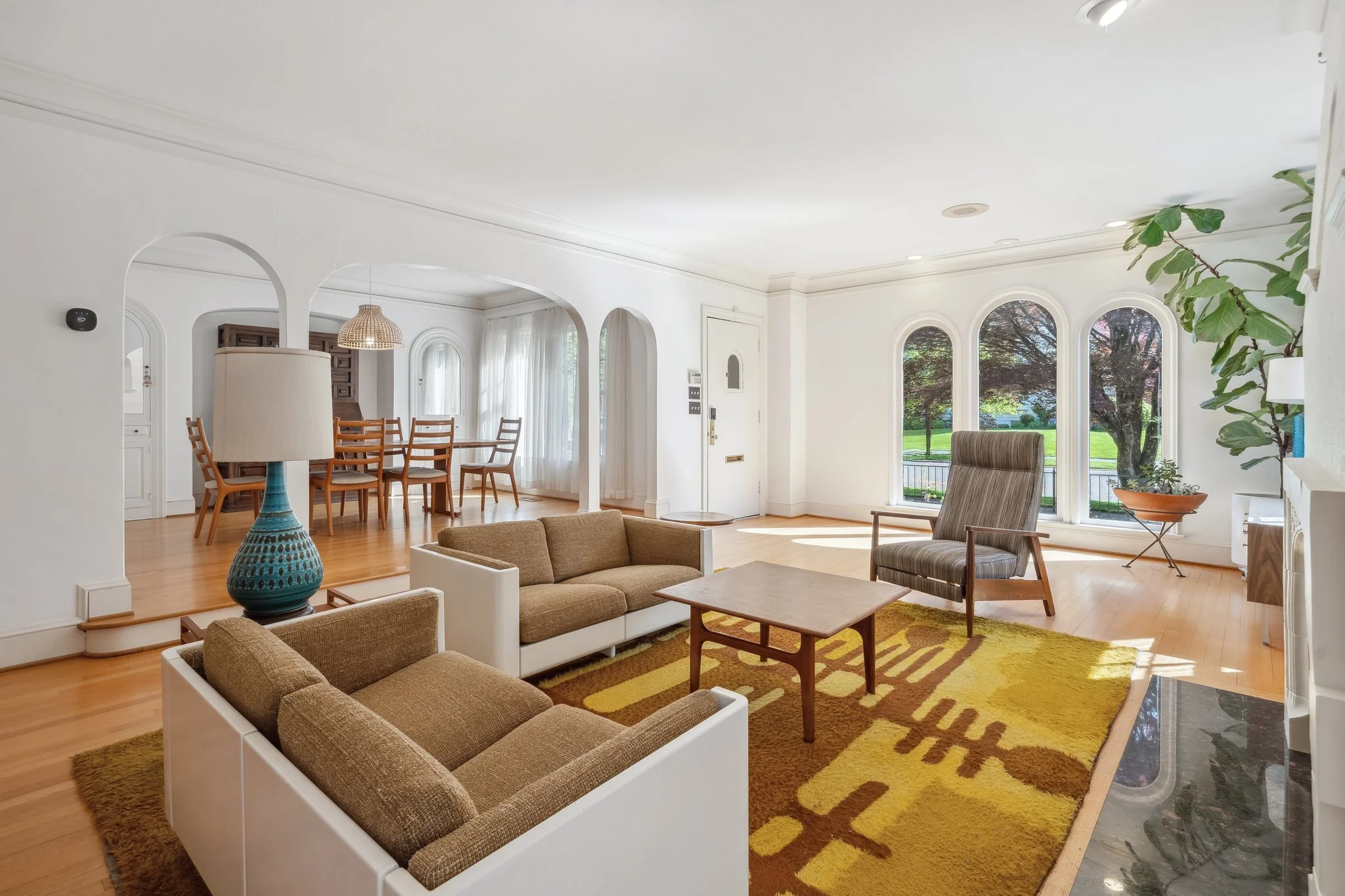 Bright living room with large windows overlooking a green yard, featuring beige and brown furniture, a yellow patterned rug, and a large green potted plant.