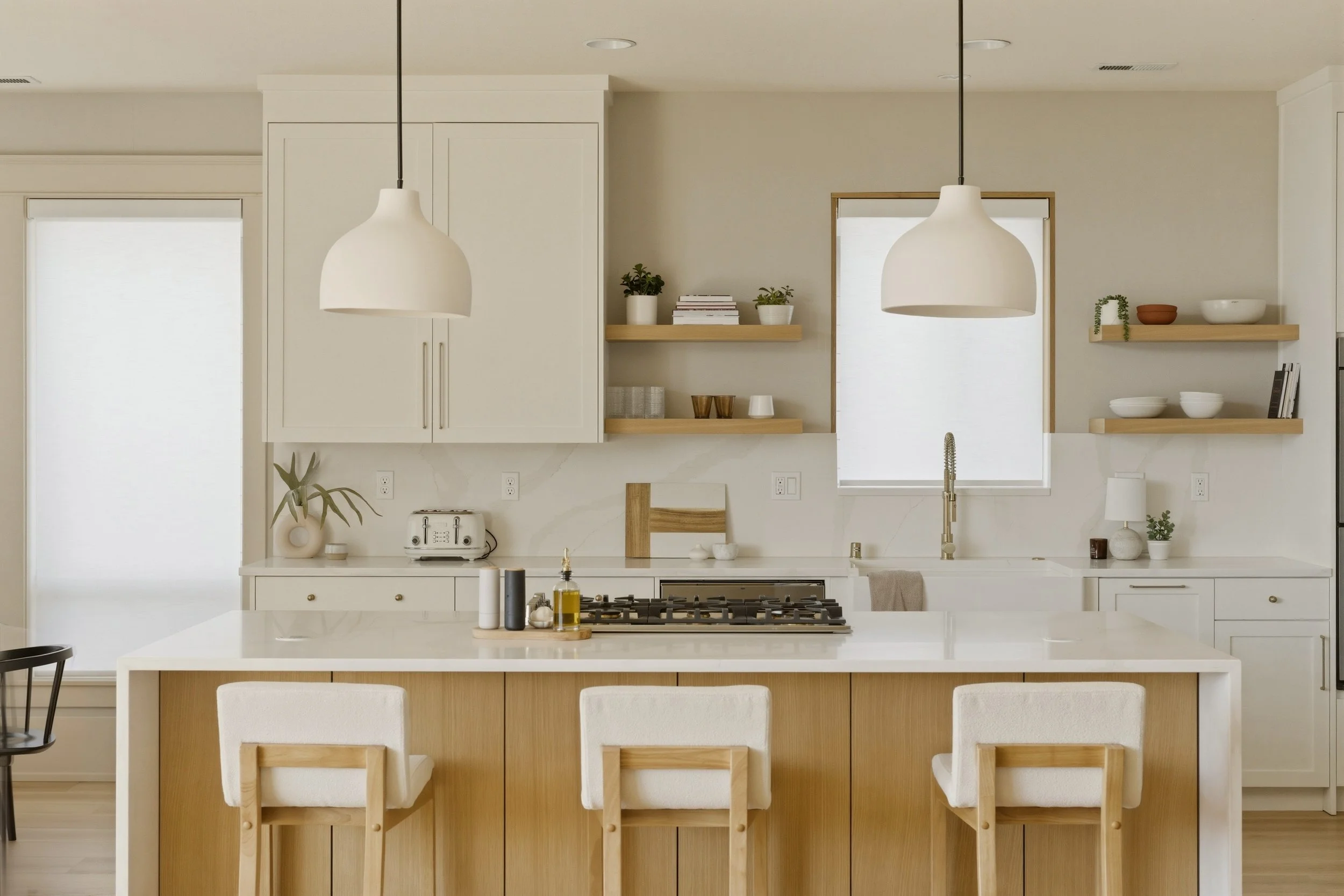 Modern kitchen with white cabinetry, open wooden shelves with bowls, plants, and books, a kitchen island with chairs, pendant lighting, a window with roller shade, and minimal decor.