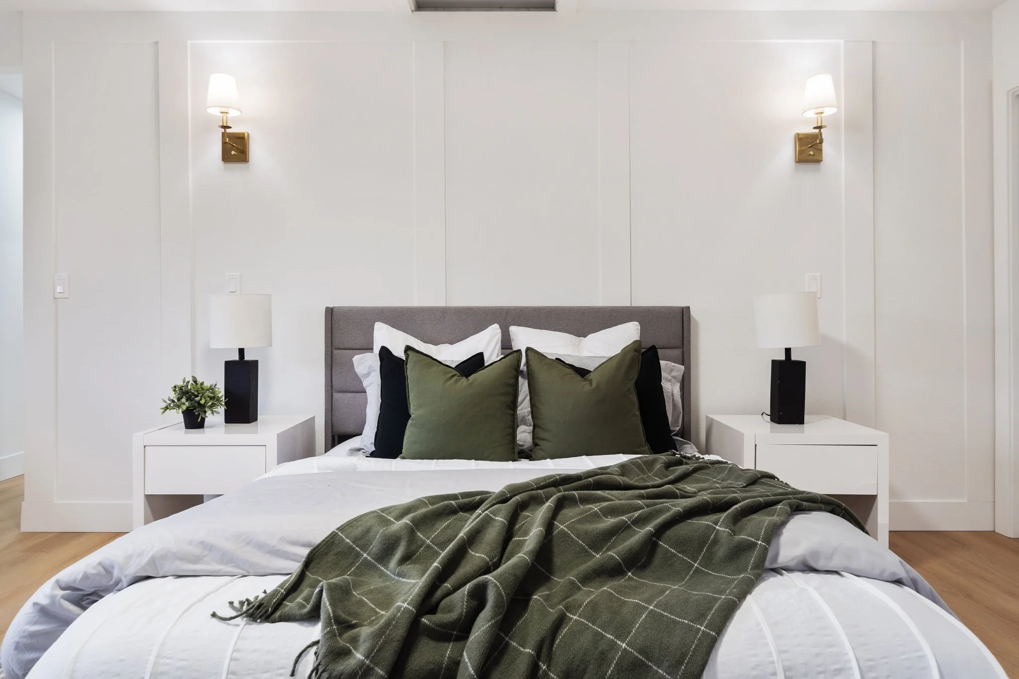 A modern bedroom featuring a bed with white bedding, a gray upholstered headboard, and multiple pillows in white, black, and green. There are two white nightstands on each side of the bed, each with a white table lamp and a small green plant. The wal