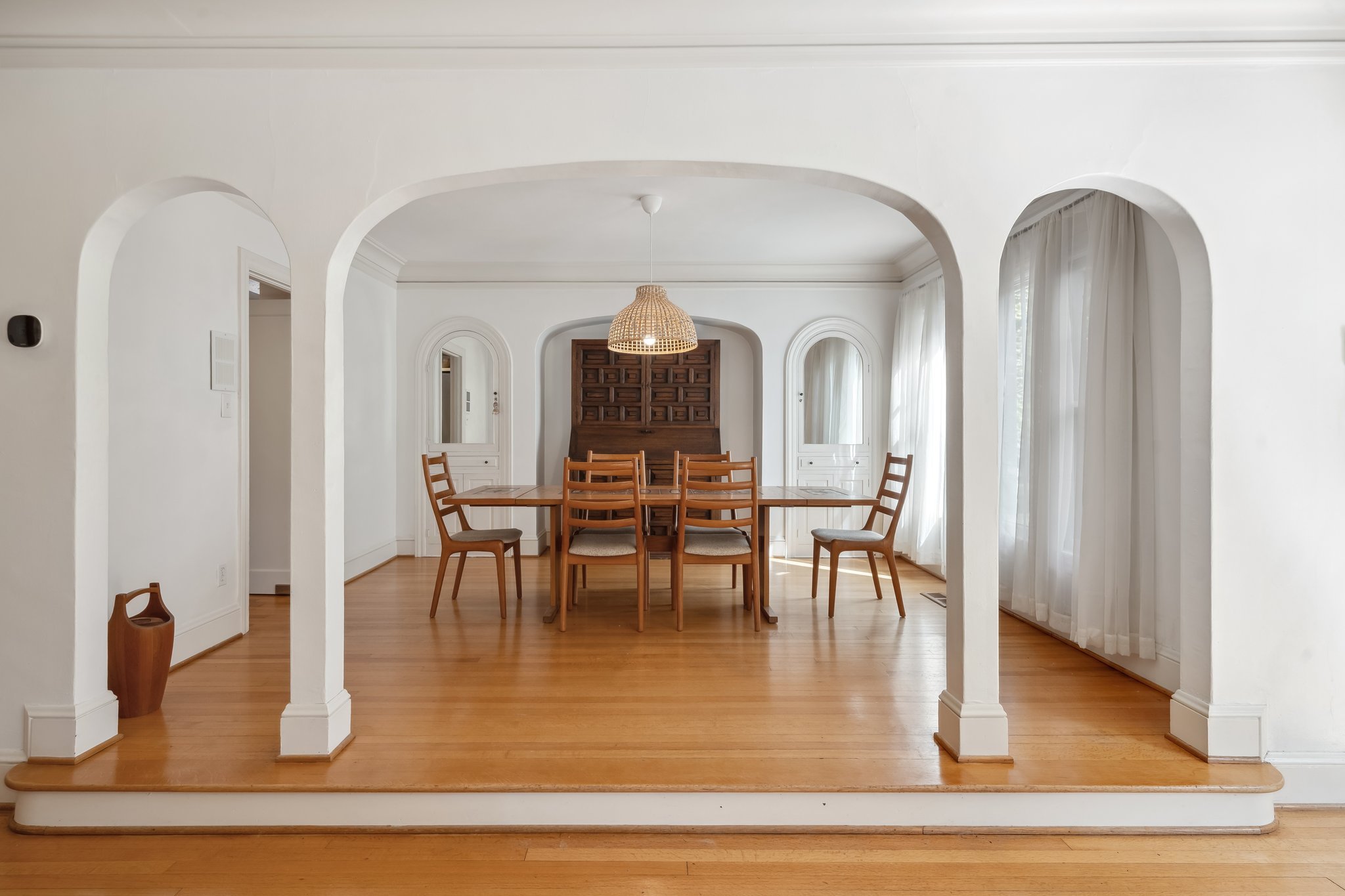 A dining room with a wooden table surrounded by six wooden chairs, a rattan pendant light hanging above, and white curtains on large windows. There are arched openings in the white walls that lead to other rooms.