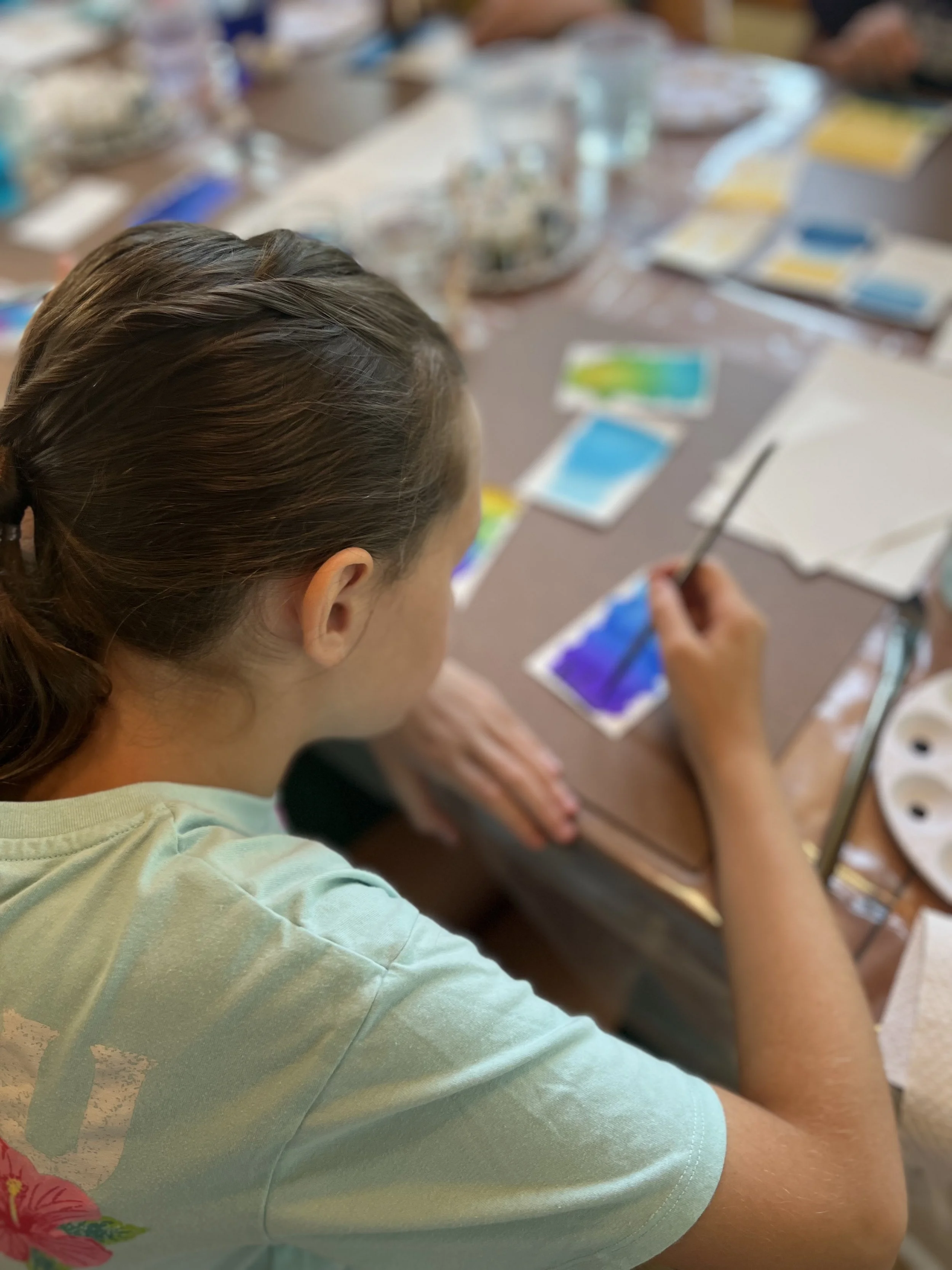 A person with long brown hair painting watercolor swatches on paper at a table, with paint supplies and other people in the background.