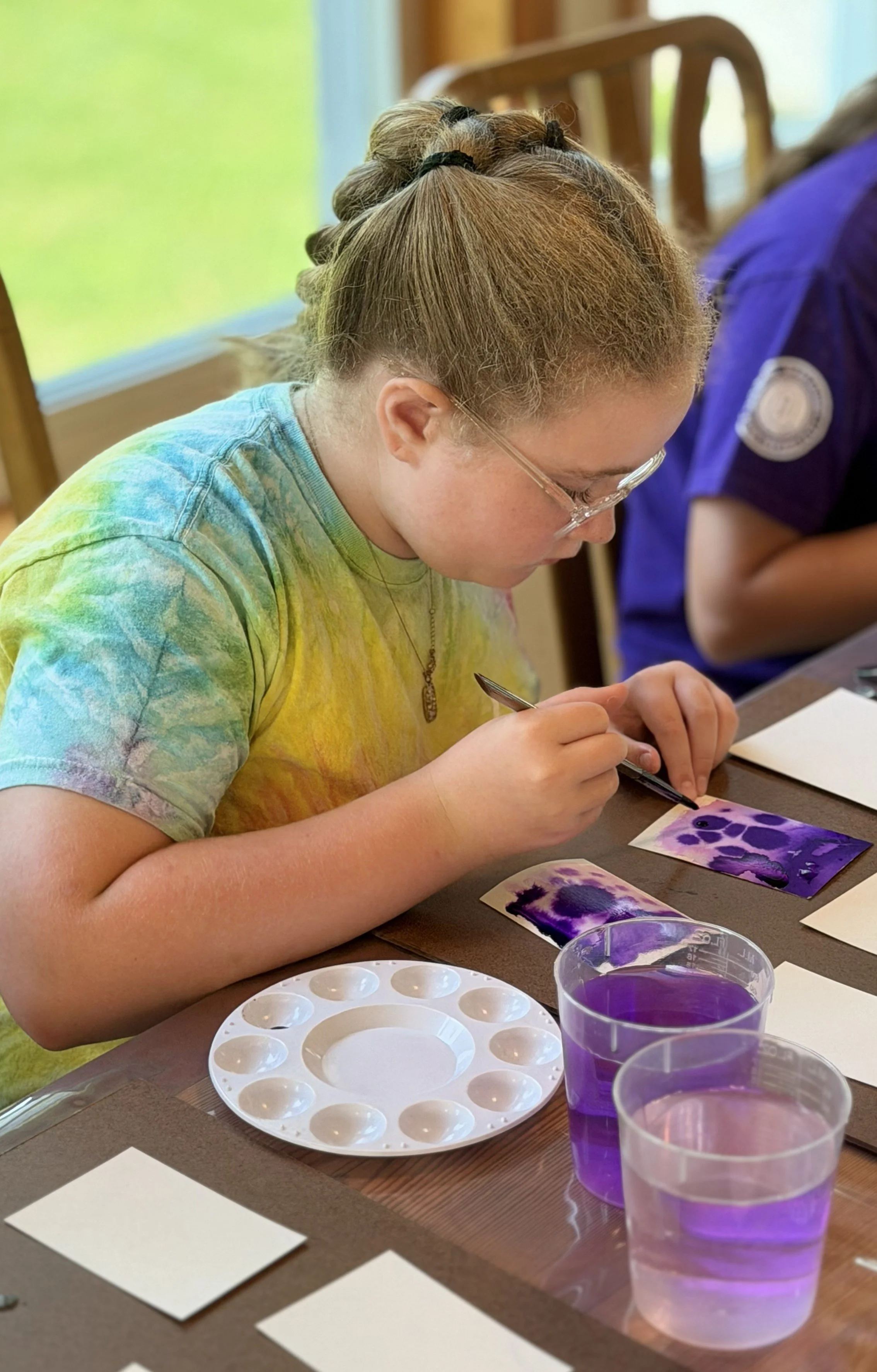 A girl with glasses and a tie-dye shirt is painting watercolor artworks in shades of purple on paper. There are paint palettes, cups of purple water, and blank cards on the table.
