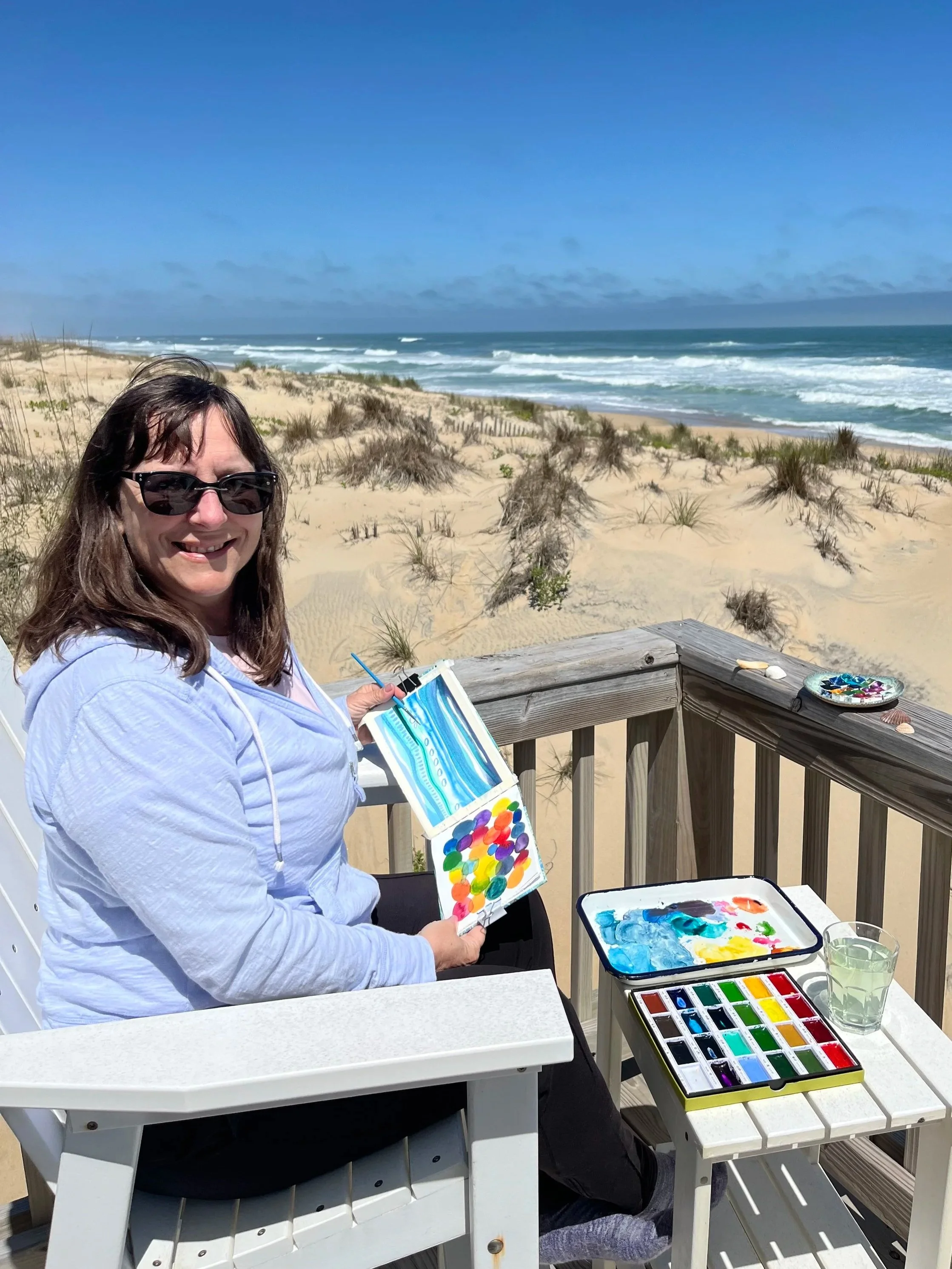 A woman with sunglasses smiling and holding a watercolor palette while sitting on a deck by the beach. Art supplies, including watercolor paints, a water container, and brushes are on the table in front of her. The beach is sandy with dunes and sparse vegetation, and the ocean with waves is visible under a blue sky.