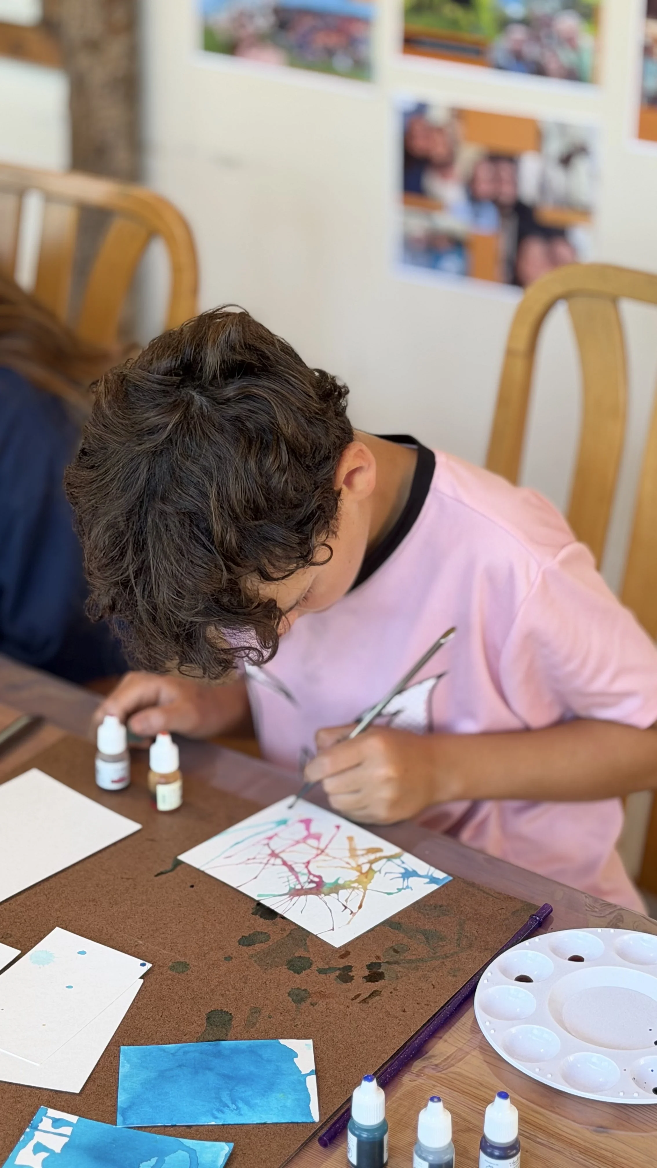 A young person with brown curly hair is painting with watercolors on a piece of paper, sitting at a table with art supplies, including bottles of paint, a water palette, and brushes, in a room with artwork on the walls.