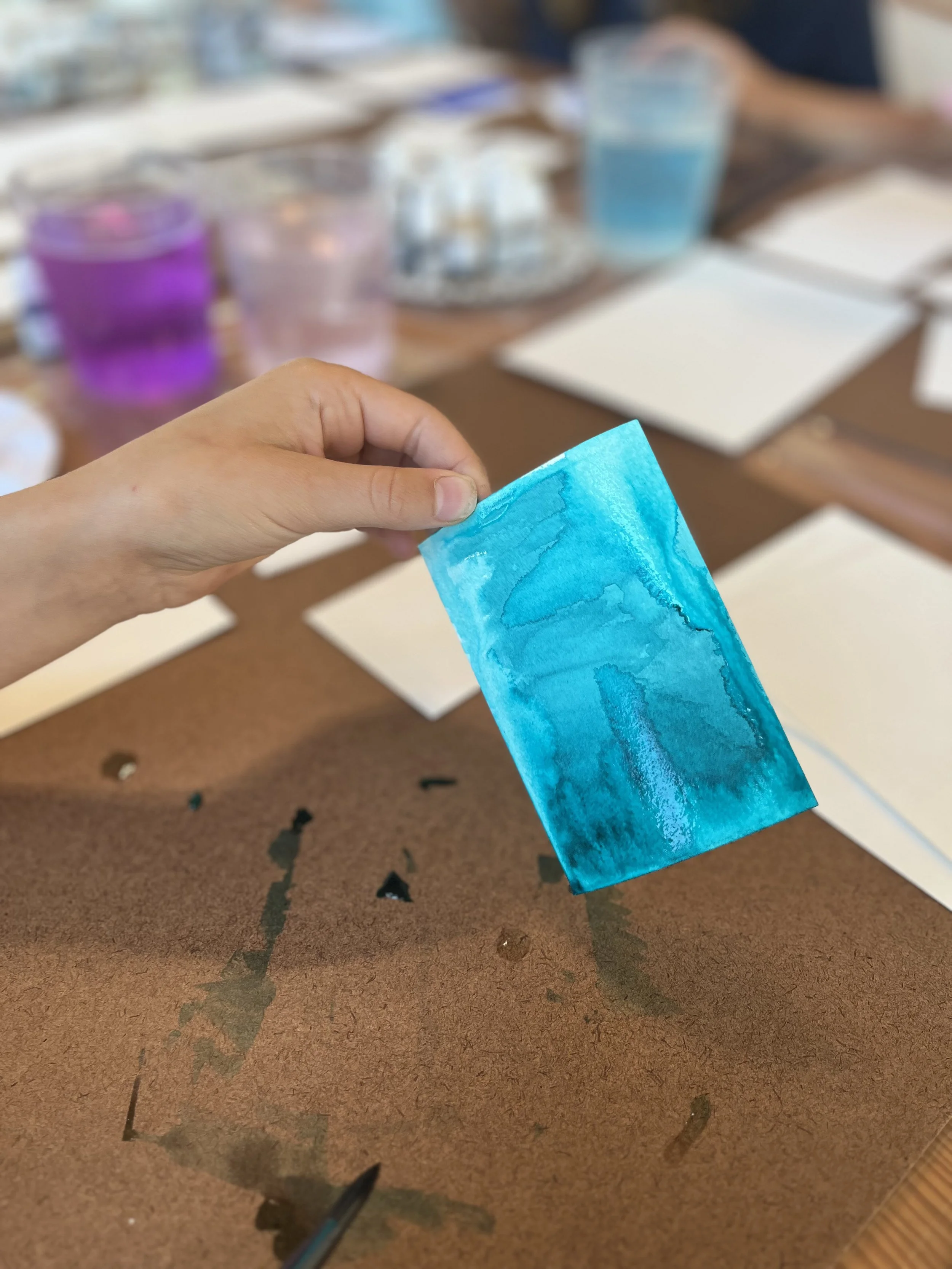 A person's hand holding a blue piece of paper with a watercolor texture. The background shows a table with blurred liquids in purple, pink, and blue glasses, along with some paper sheets and art supplies.