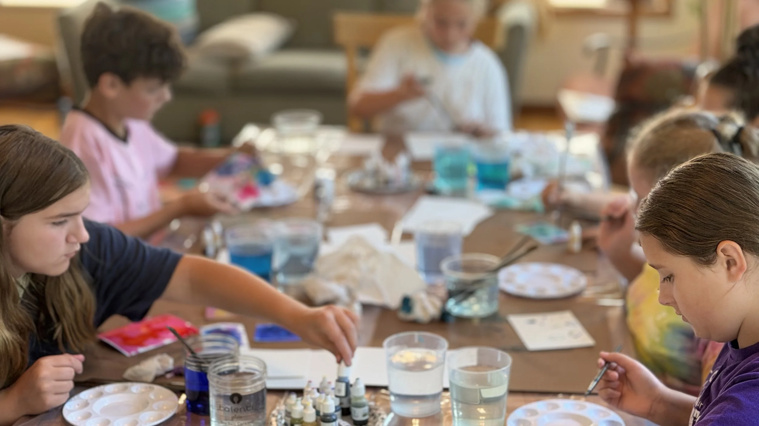Children engaged in an art or science activity at a table with various supplies, supervised by an adult in a well-lit room.