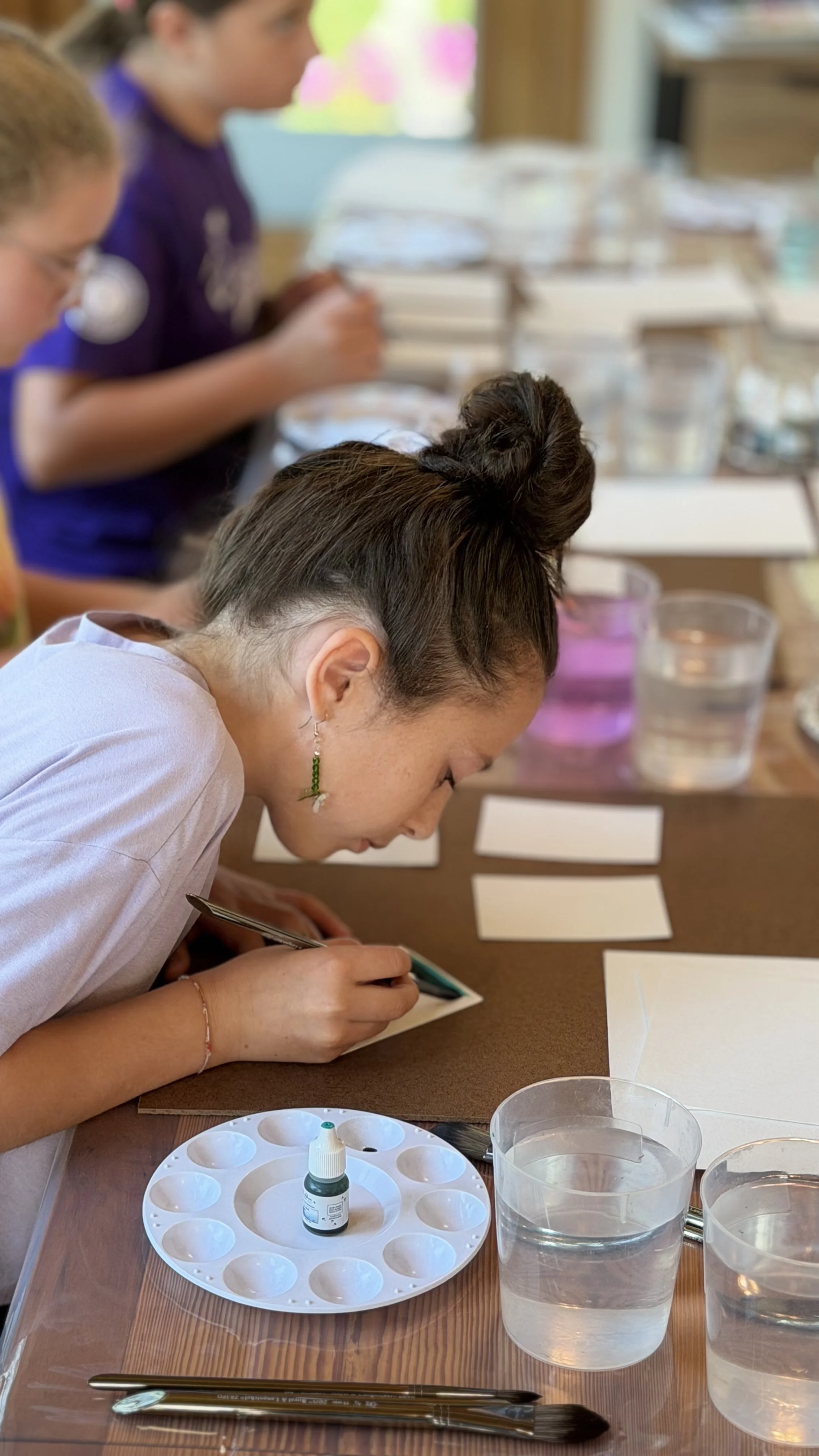 A woman with earrings and a buns hairstyle is painting with watercolors or ink on paper at a table, with her eyes focused and her hands holding a small brush or pen. There are small pots for mixing colors, a paint palette with water, and several glasses of water on the table, indicating an art or craft activity.