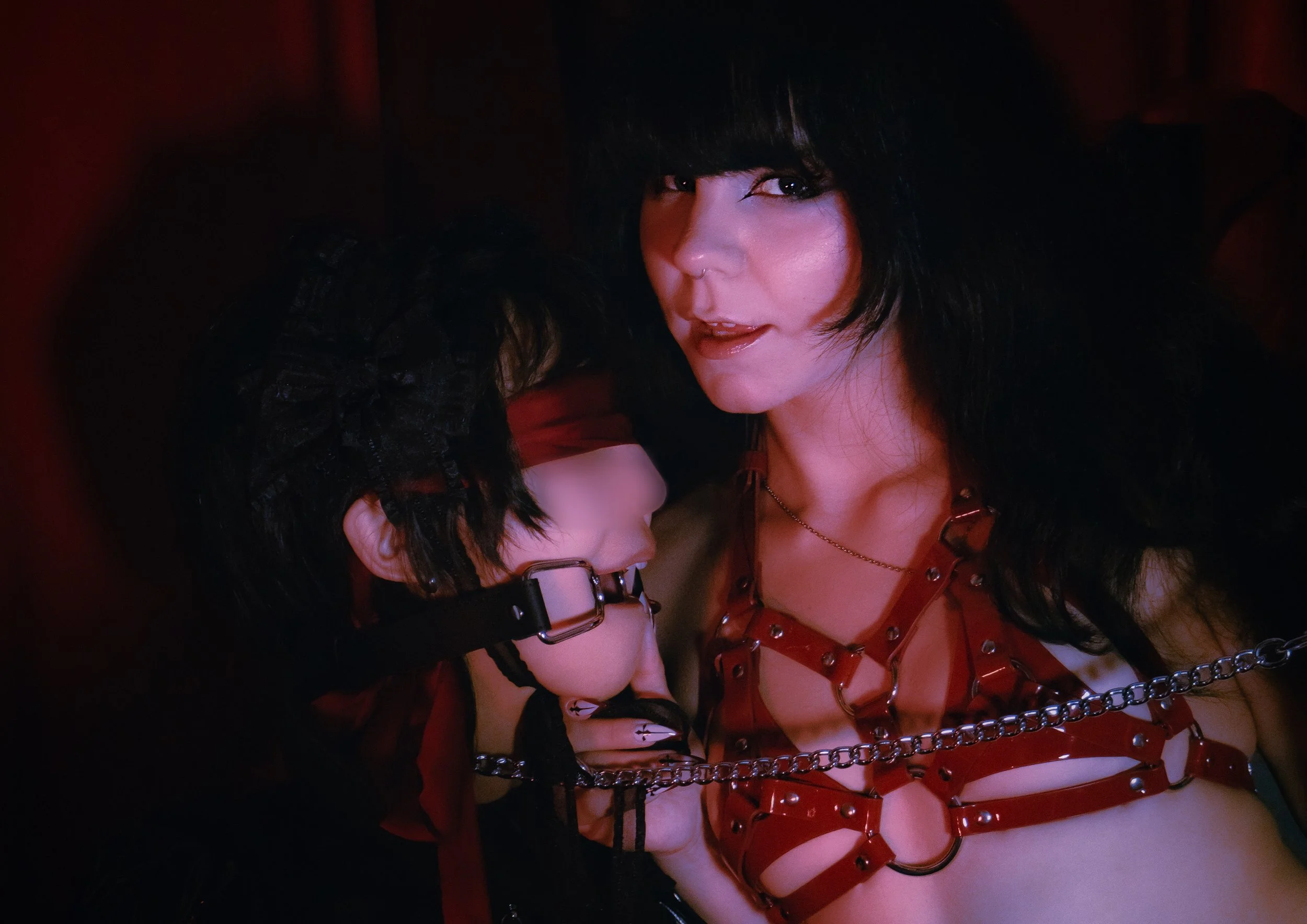 A woman with dark hair and makeup, wearing a red outfit with metal chains and straps, and a man with glasses and dark hair, in a close embrace in a dark, intimate setting.
