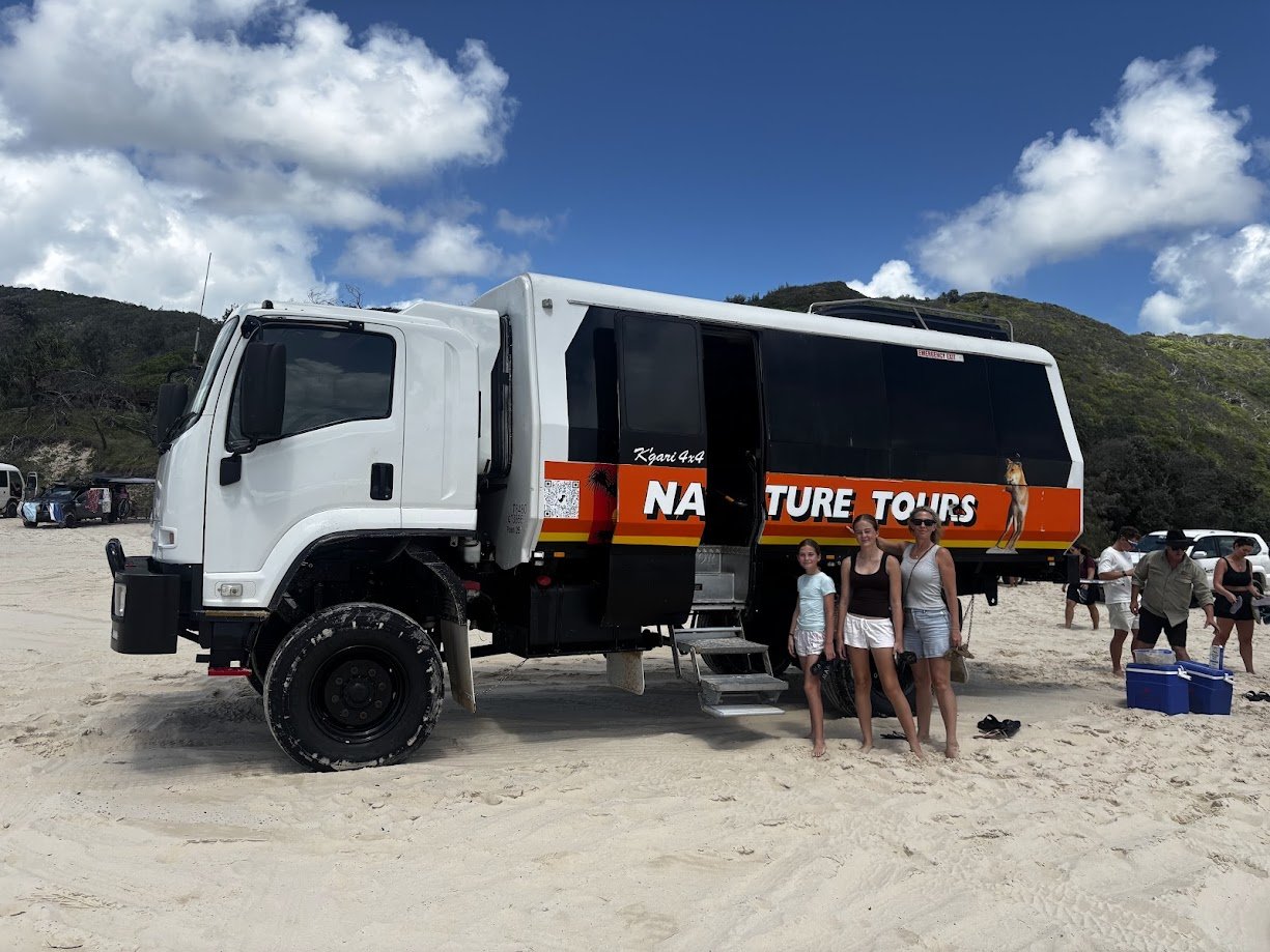 A white truck with a black and orange 'Nature Tours' sign parked on a sandy beach, with three women standing in front of it, and other people in the background near trees and vehicles under a partly cloudy sky.