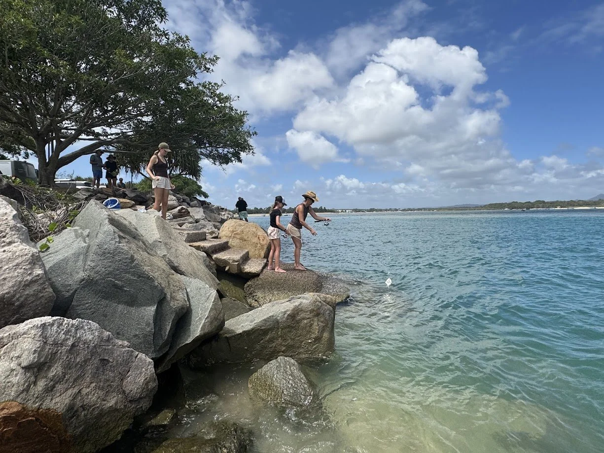 People fishing off rocks by the bay on a partly cloudy day, with trees and distant shoreline visible.