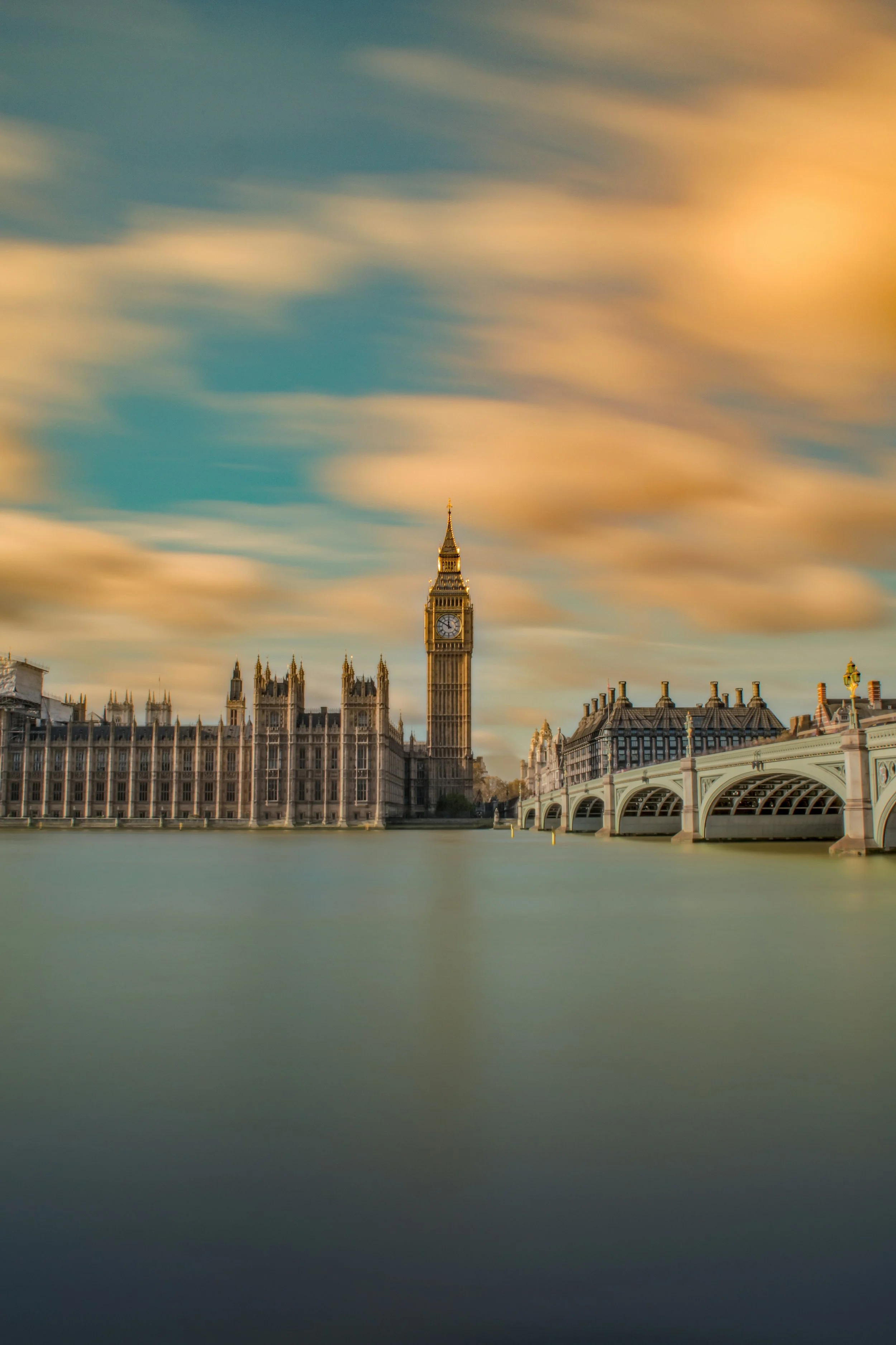 Big Ben clock tower and the Palace of Westminster in London, England, during sunset with moving clouds and a calm river in the foreground.