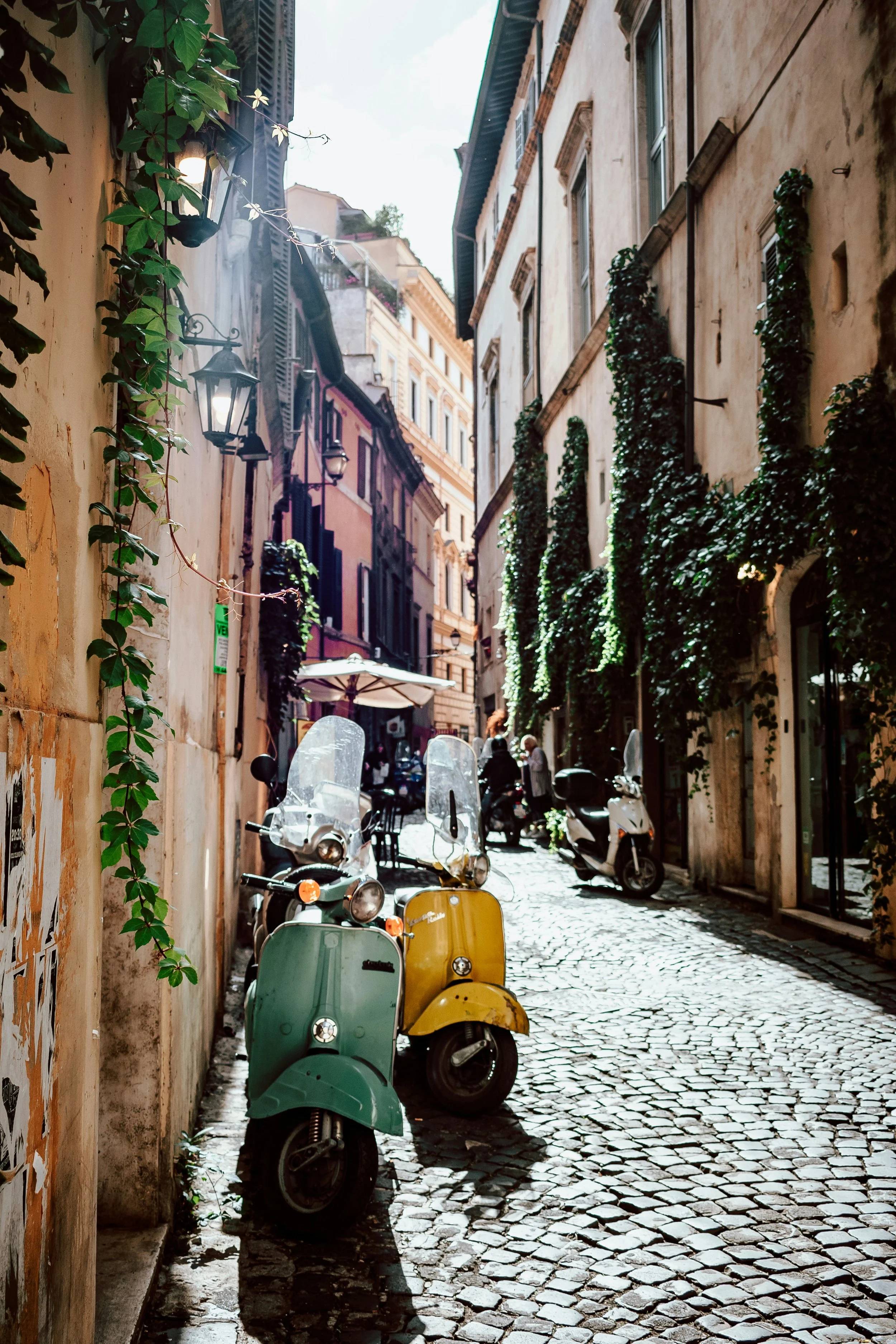 A narrow cobblestone street lined with old buildings, with green and yellow scooters parked along the side and greenery climbing on the walls.
