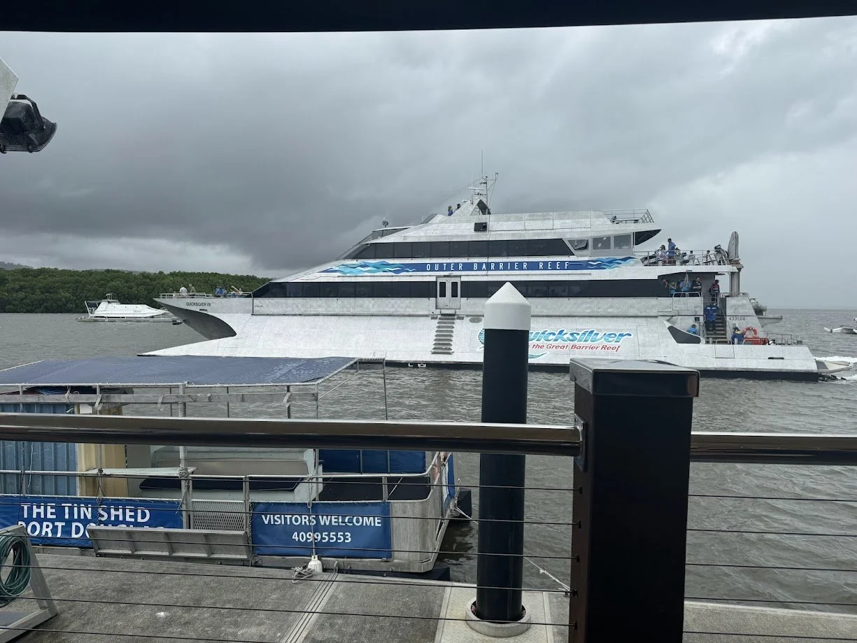 A large boat labeled 'Quicksilver' with 'Outer Barrier Reef' written above it is docked at a pier on a cloudy day, with smaller boats nearby.
