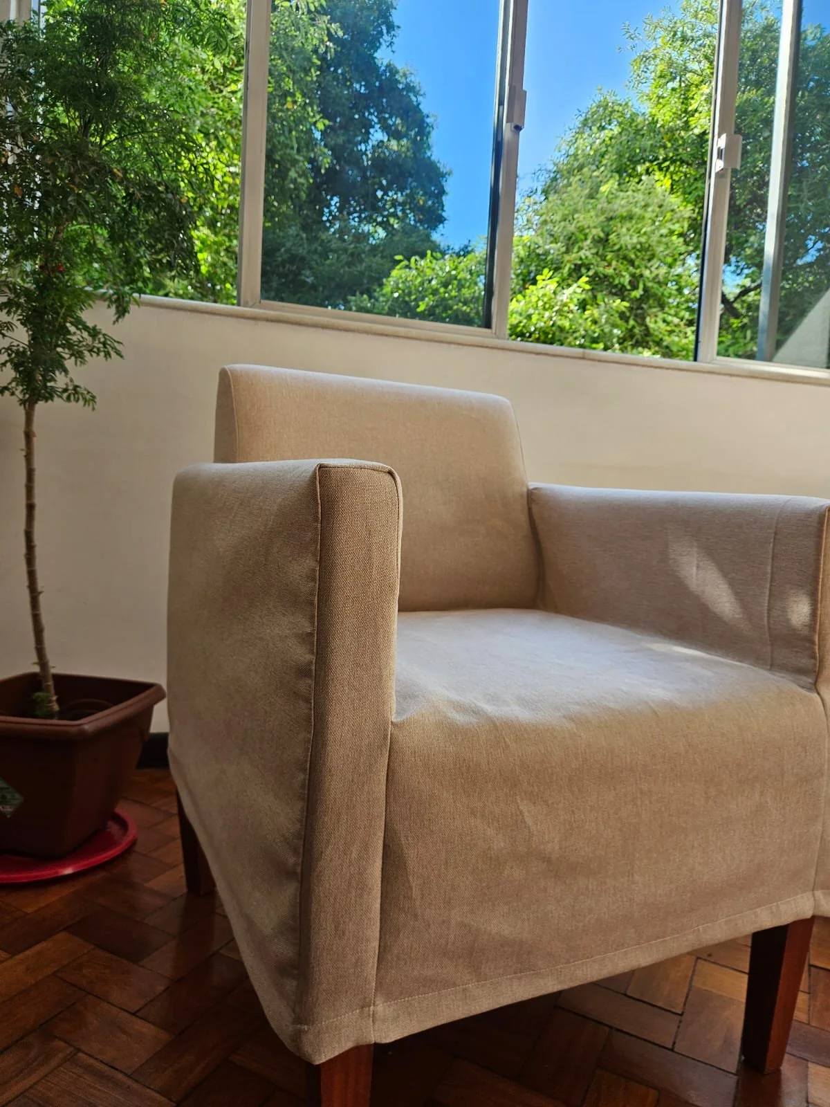 Poltrona bege de tecido em ambiente com janela grande, vidro aberto, árvores verdes e céu azul ao fundo, em um cômodo com piso de madeira.